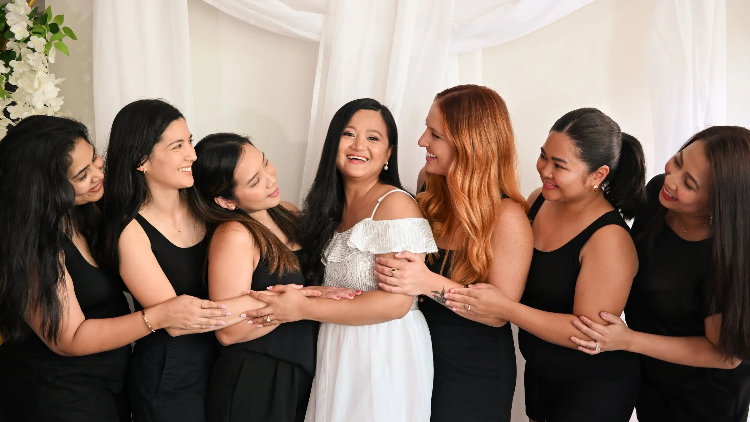 The same moment in a brighter edit: Winnie laughing in her white dress as her bridesmaids in black link arms around her