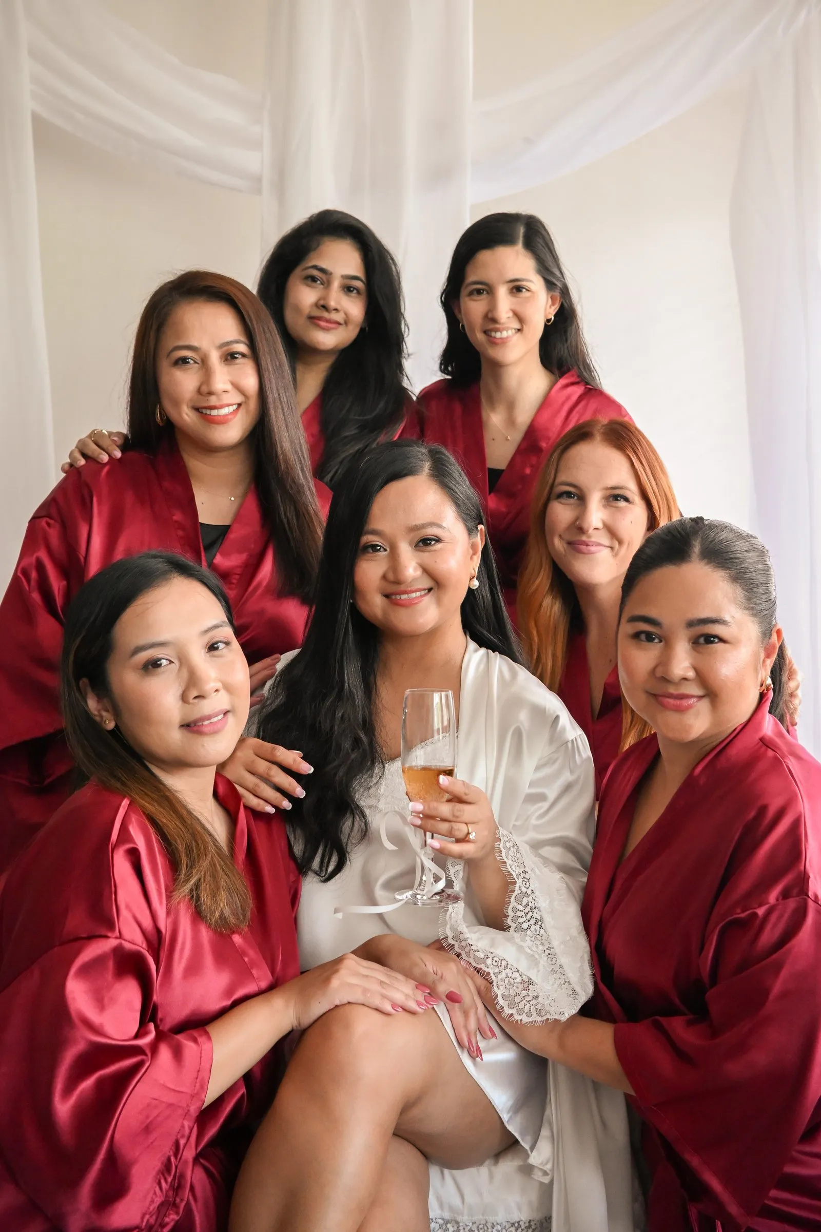 Winnie holding a glass of champagne while her bridesmaids in red robes gather close behind her