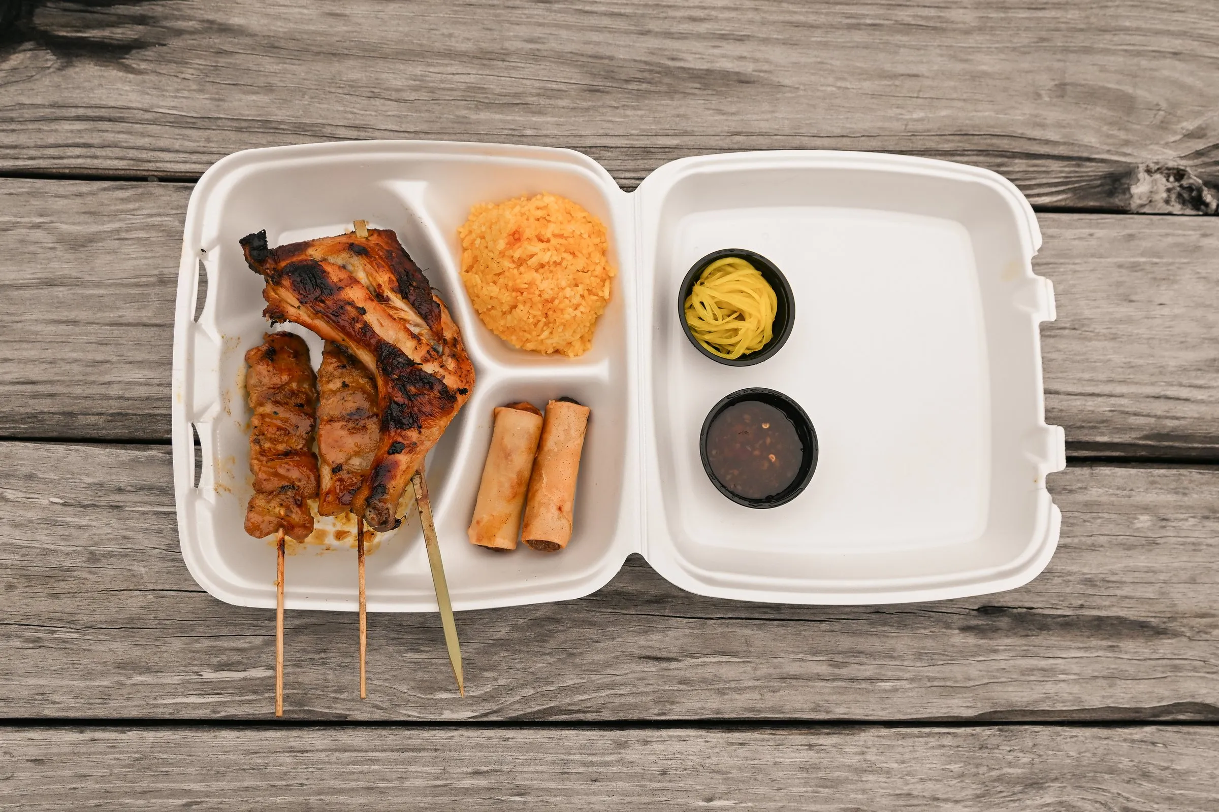 Overhead view of a takeout combo with chicken inasal, pork barbecue skewers, java rice, lumpia, and two dipping sauces on a picnic table