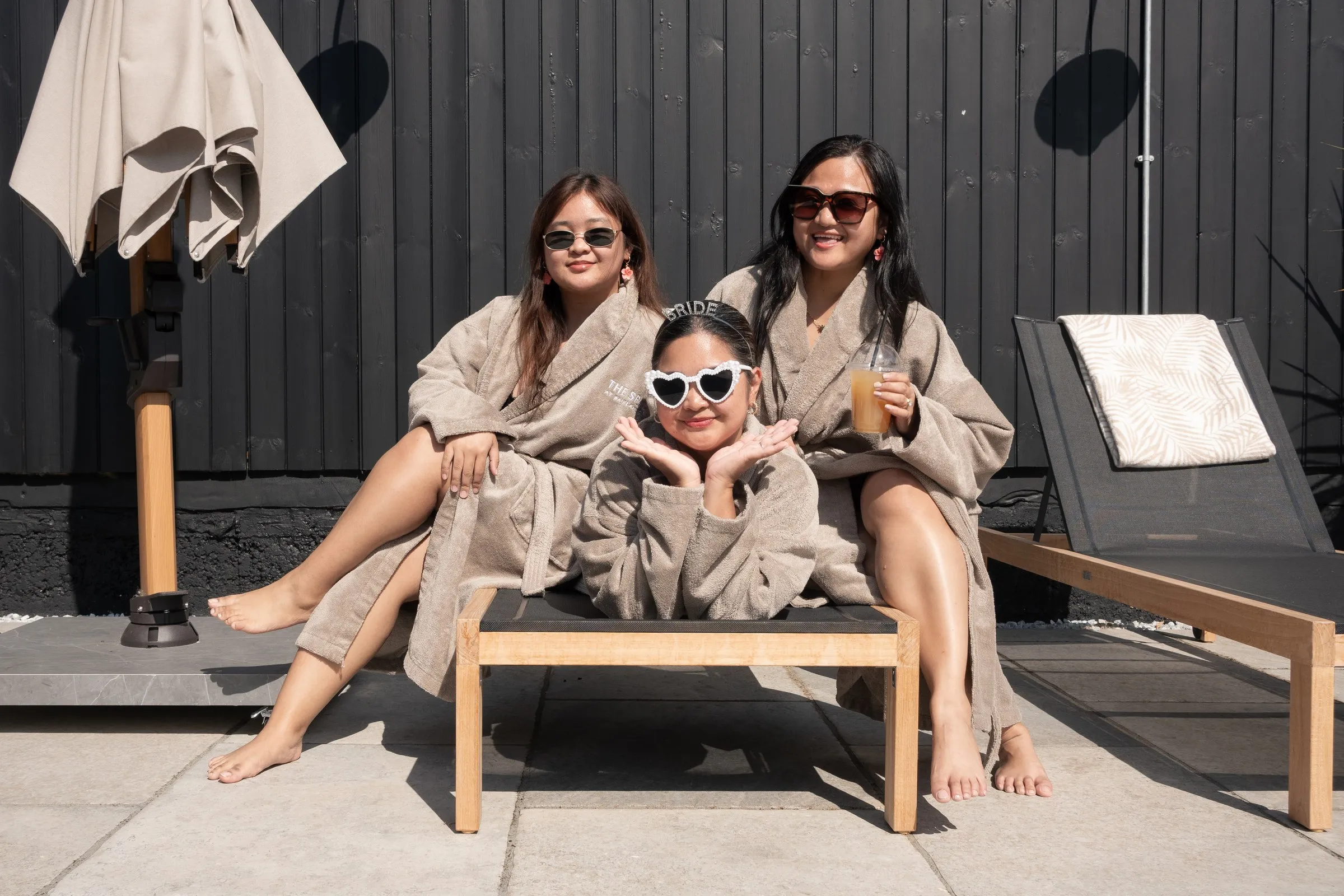 Three friends in robes and sunglasses posing together on a sun lounger