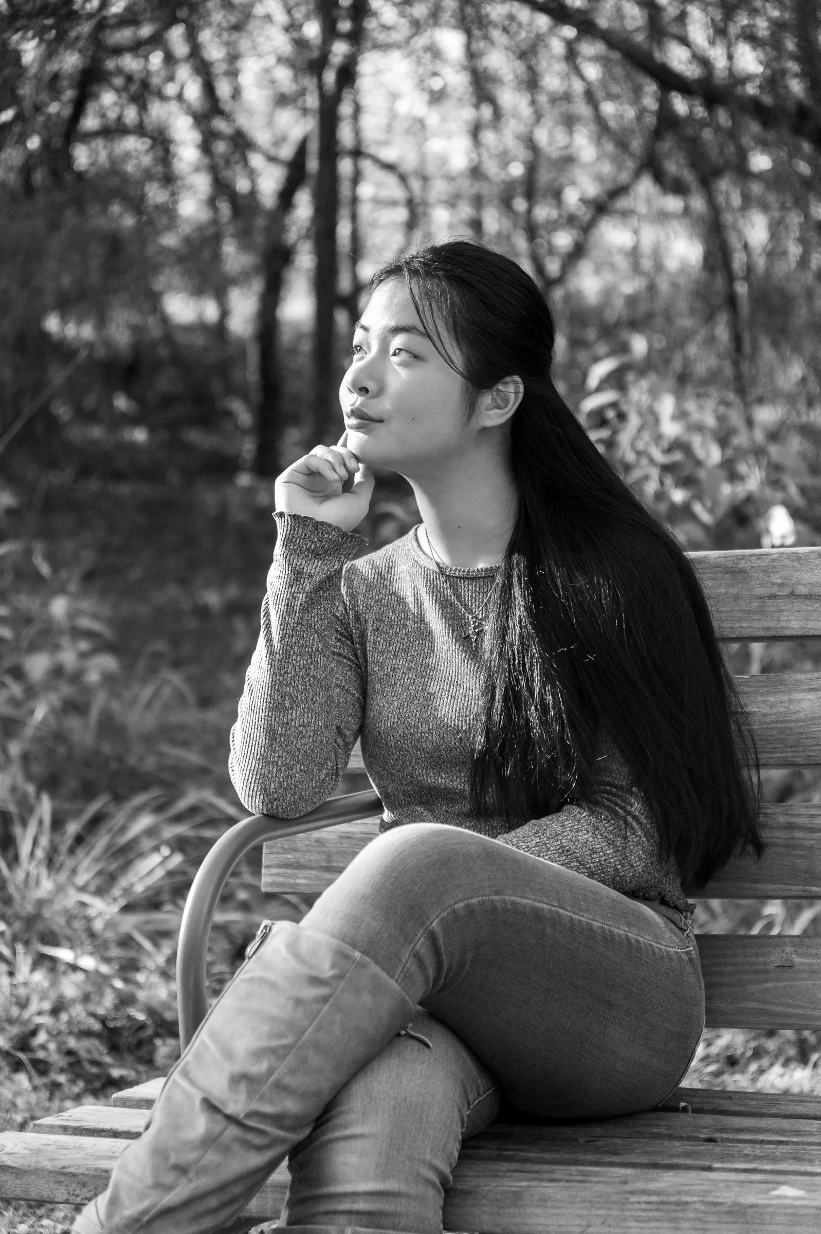 Black and white portrait of Meilin seated on a wooden park bench, chin resting on her hand as she looks off into the trees