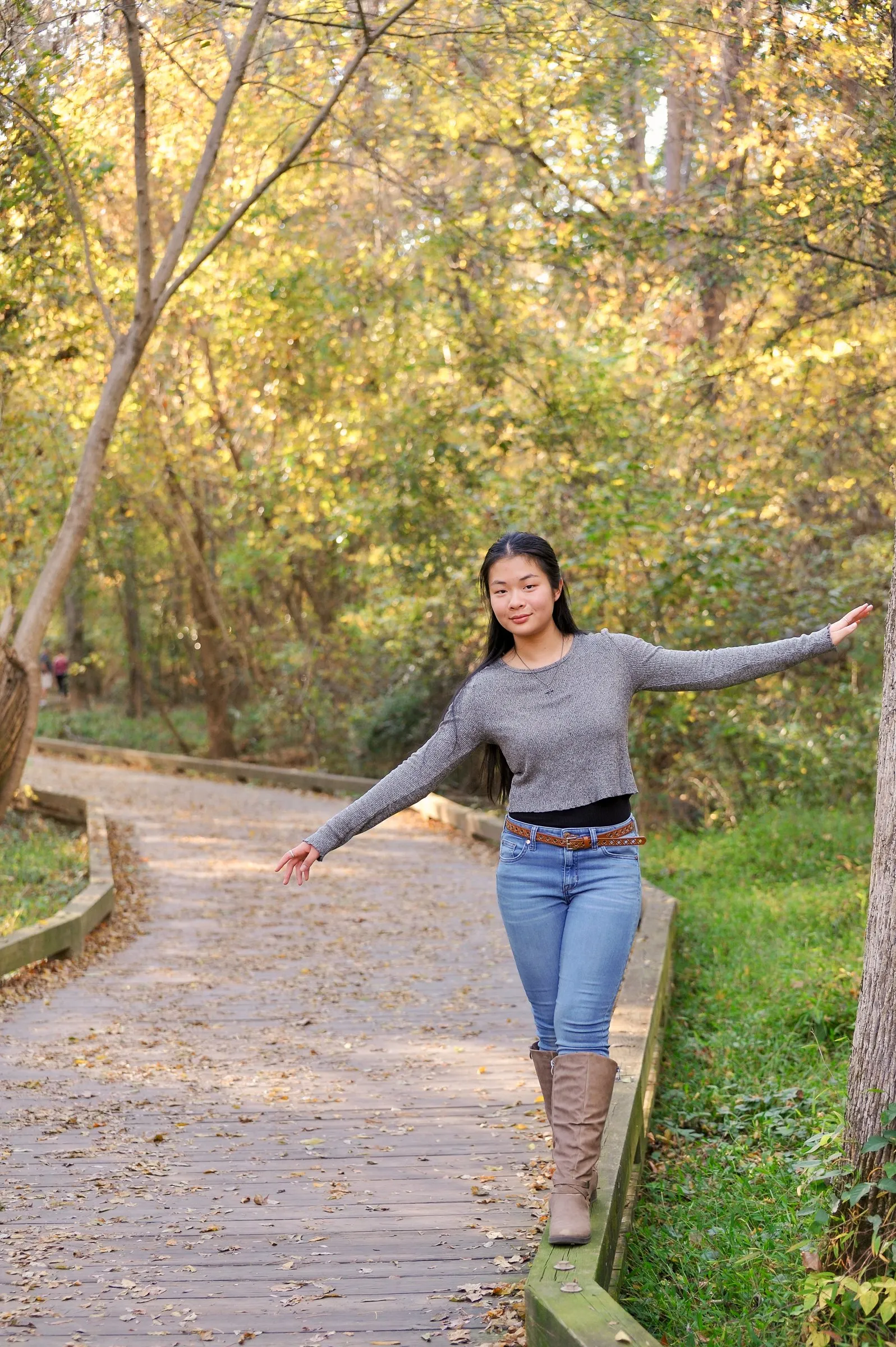 Meilin balancing along the edge of a curving boardwalk trail, arms outstretched, surrounded by fall color