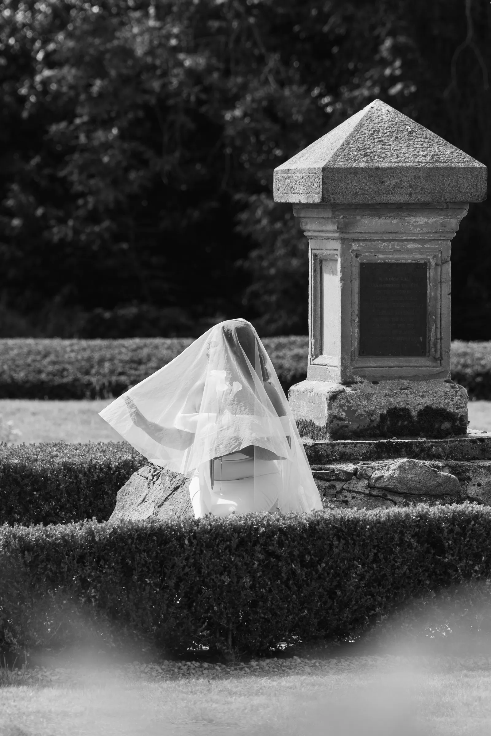 Black and white photograph of Mathelle sitting quietly beneath her veil beside a stone pillar at Rothley Court