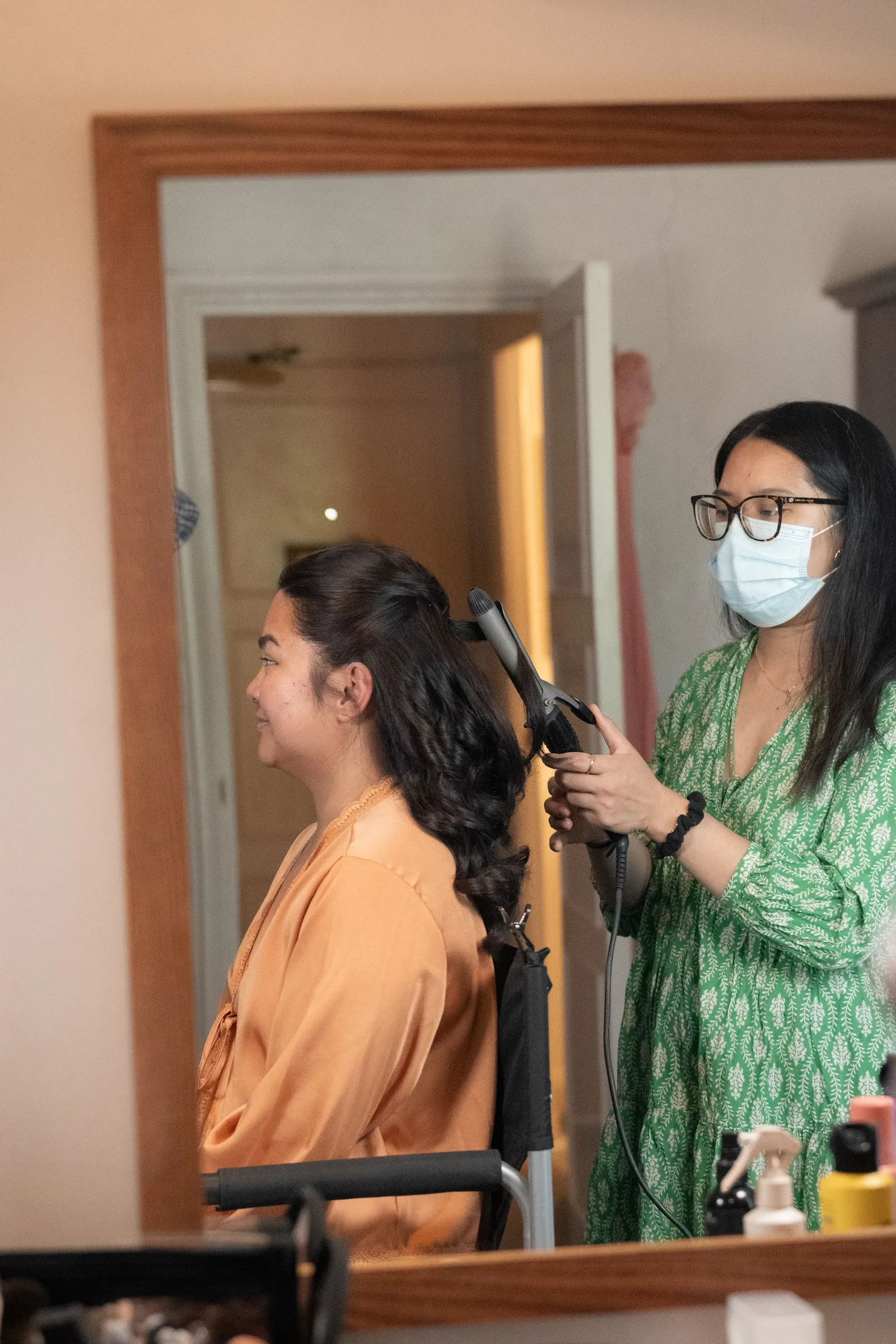 Hairstylist in a green dress curling the hair of a woman in a peach robe in the bridal suite