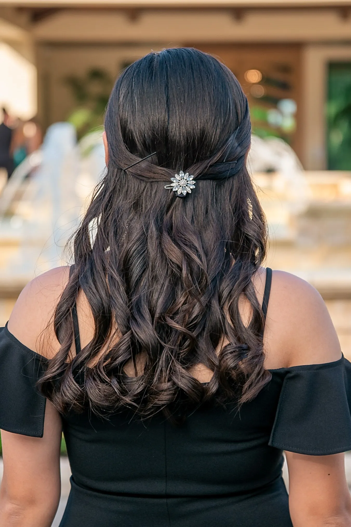 Mya from behind in an off the shoulder black gown, her long curls pinned back with a jeweled clip, fountains blurred beyond her