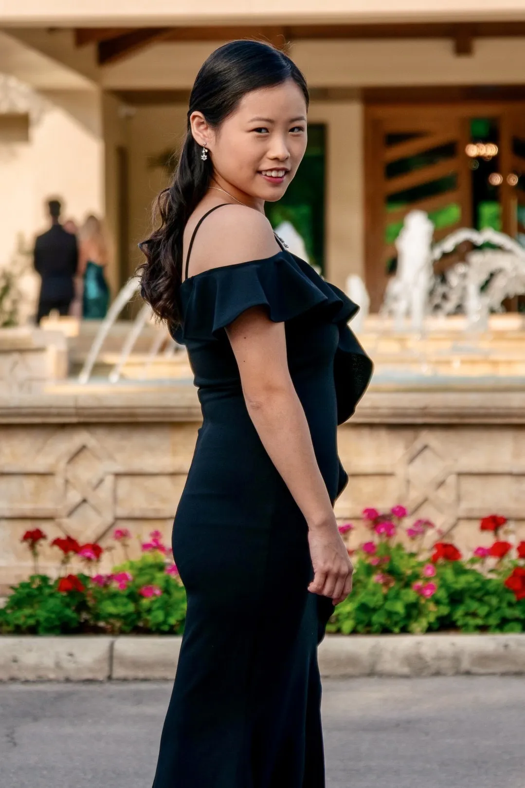 Full length portrait of Mya in a fitted black off the shoulder gown, glancing back over her shoulder in front of a fountain and red flowers