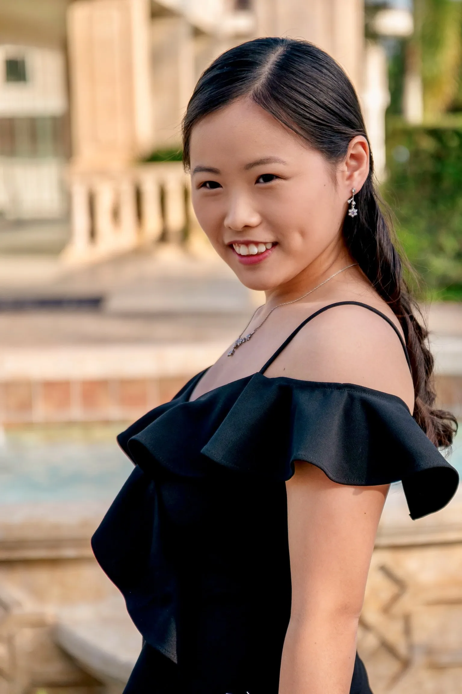 Mya smiling over her shoulder in a ruffled black gown beside a stone fountain