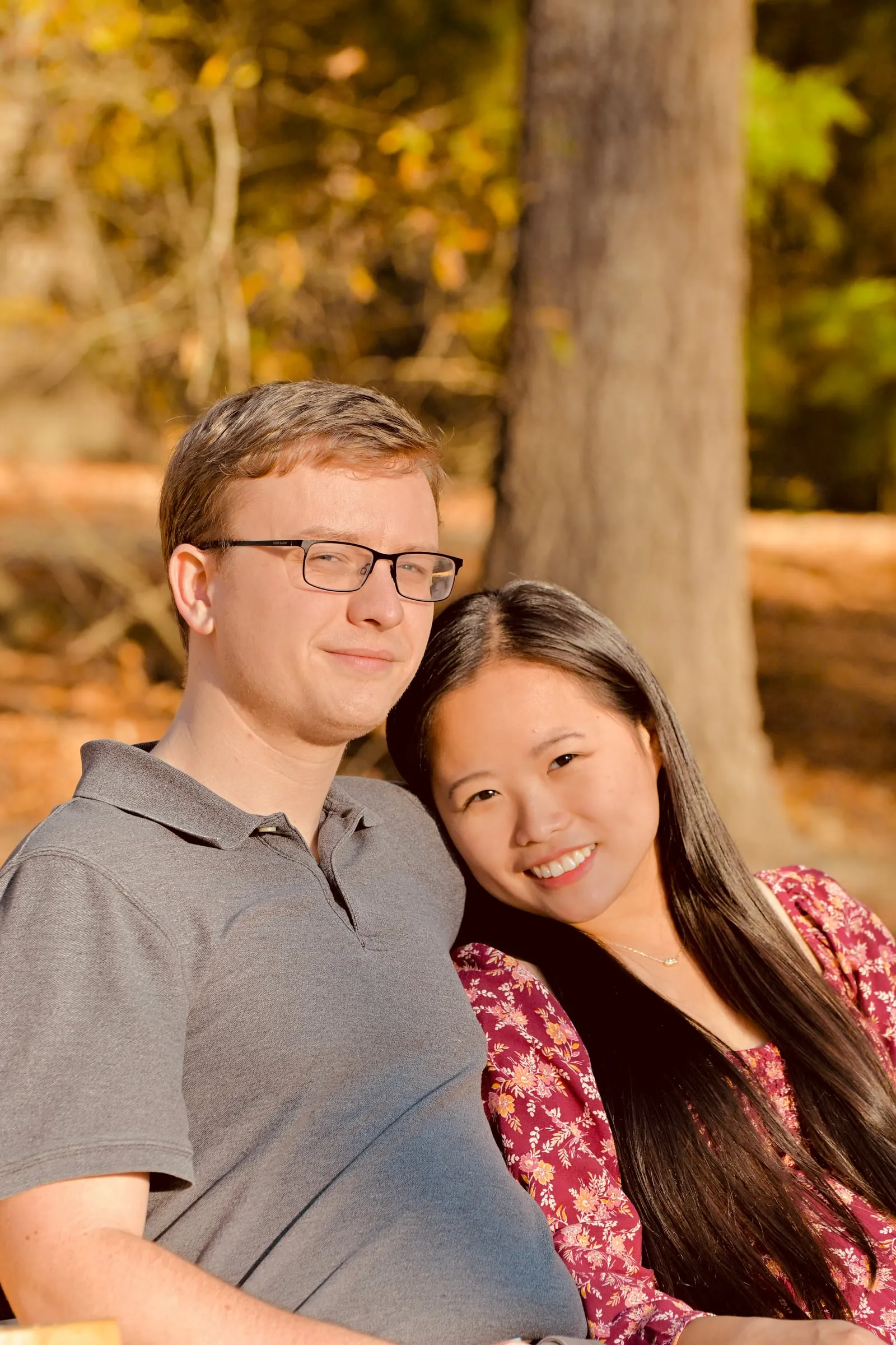 Mya resting her head on Jacob's shoulder beneath a tall tree in warm afternoon light