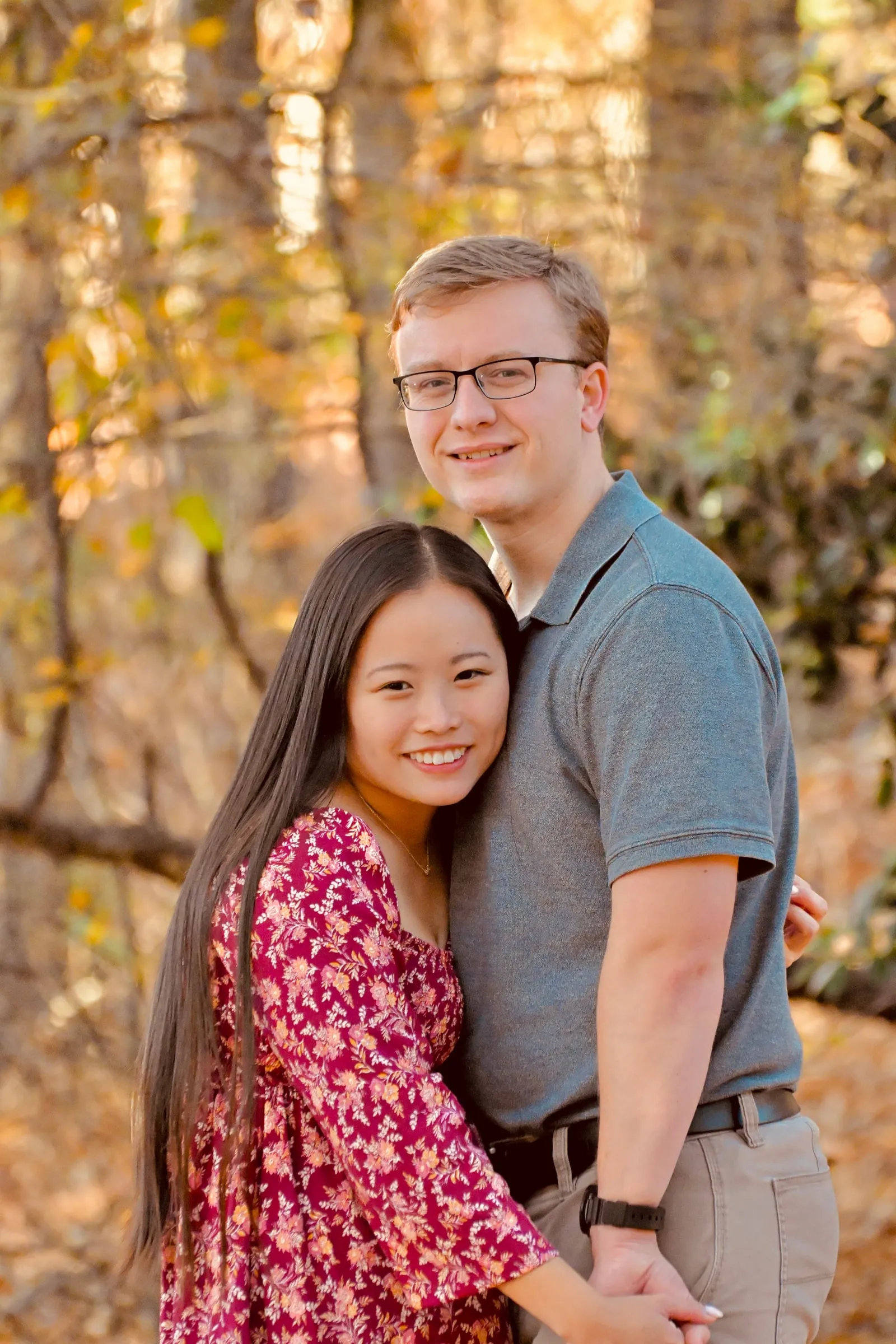 Mya and Jacob standing close and holding hands, smiling at the camera among autumn trees