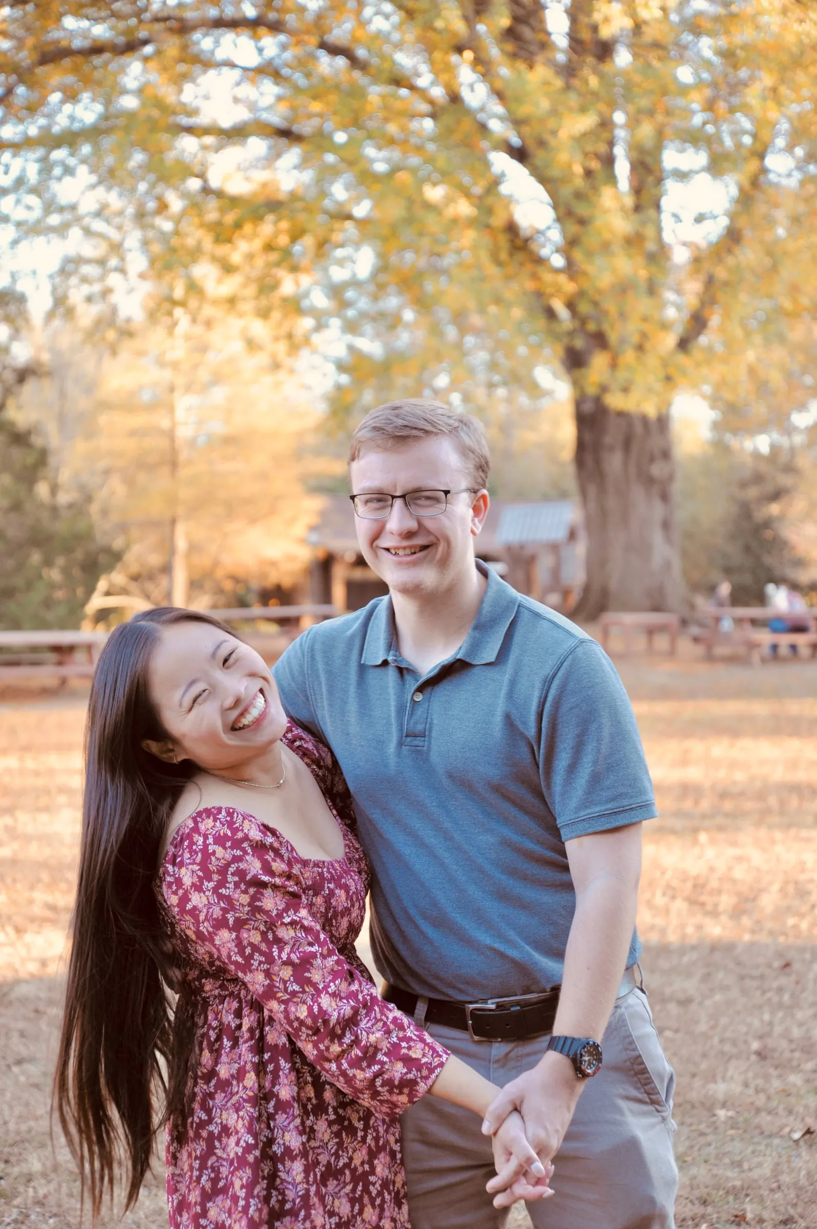 Mya laughing as she holds Jacob's hands on the open lawn, picnic shelters and a large oak behind them