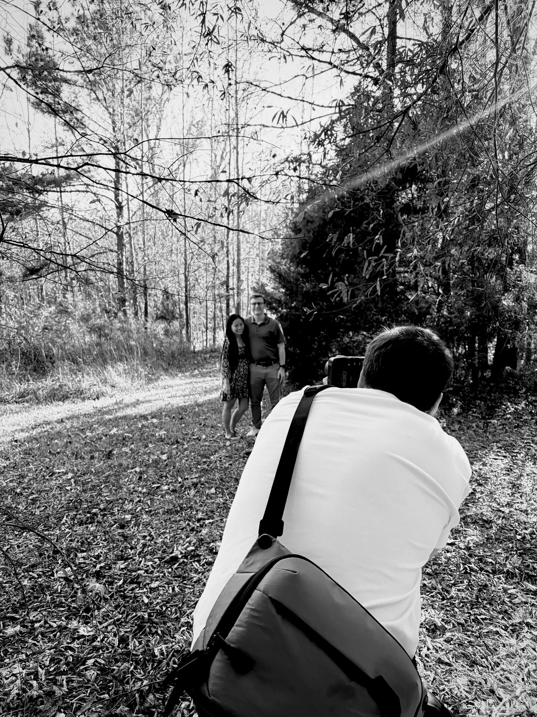 Black and white behind the scenes frame of the photographer kneeling on the leaf-covered trail, framing Mya and Jacob