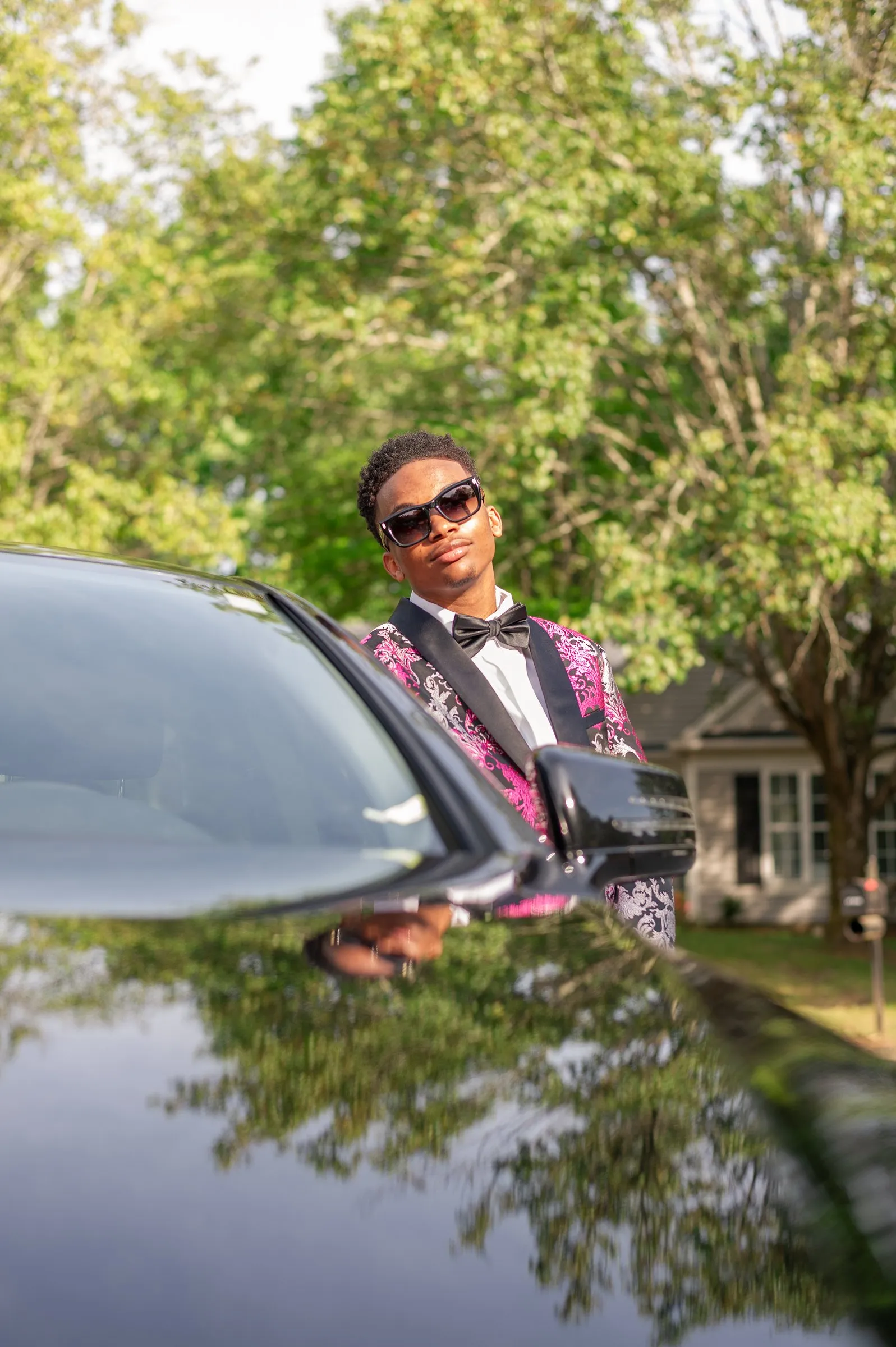 His reflection in the polished hood of a black car, trees overhead