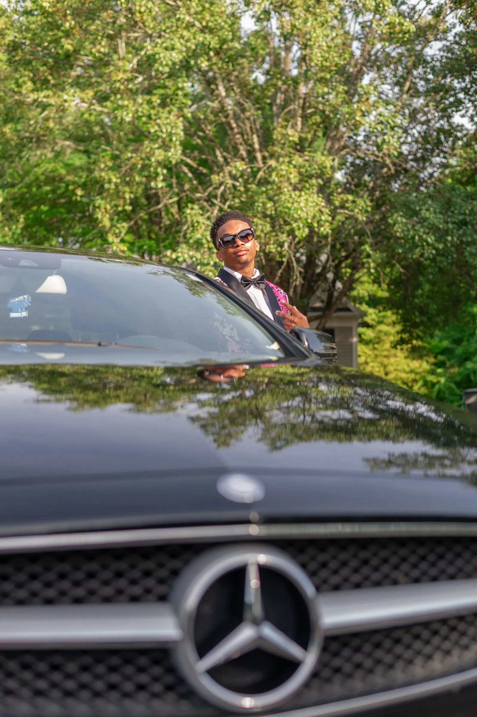 Low angle over the Mercedes grille, with him leaning against the car behind it
