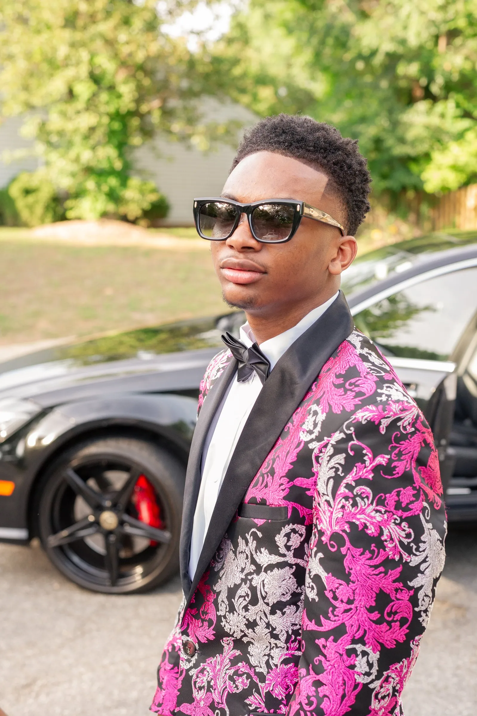 Close prom portrait in sunglasses and a black bow tie, a black car with red brake calipers behind him
