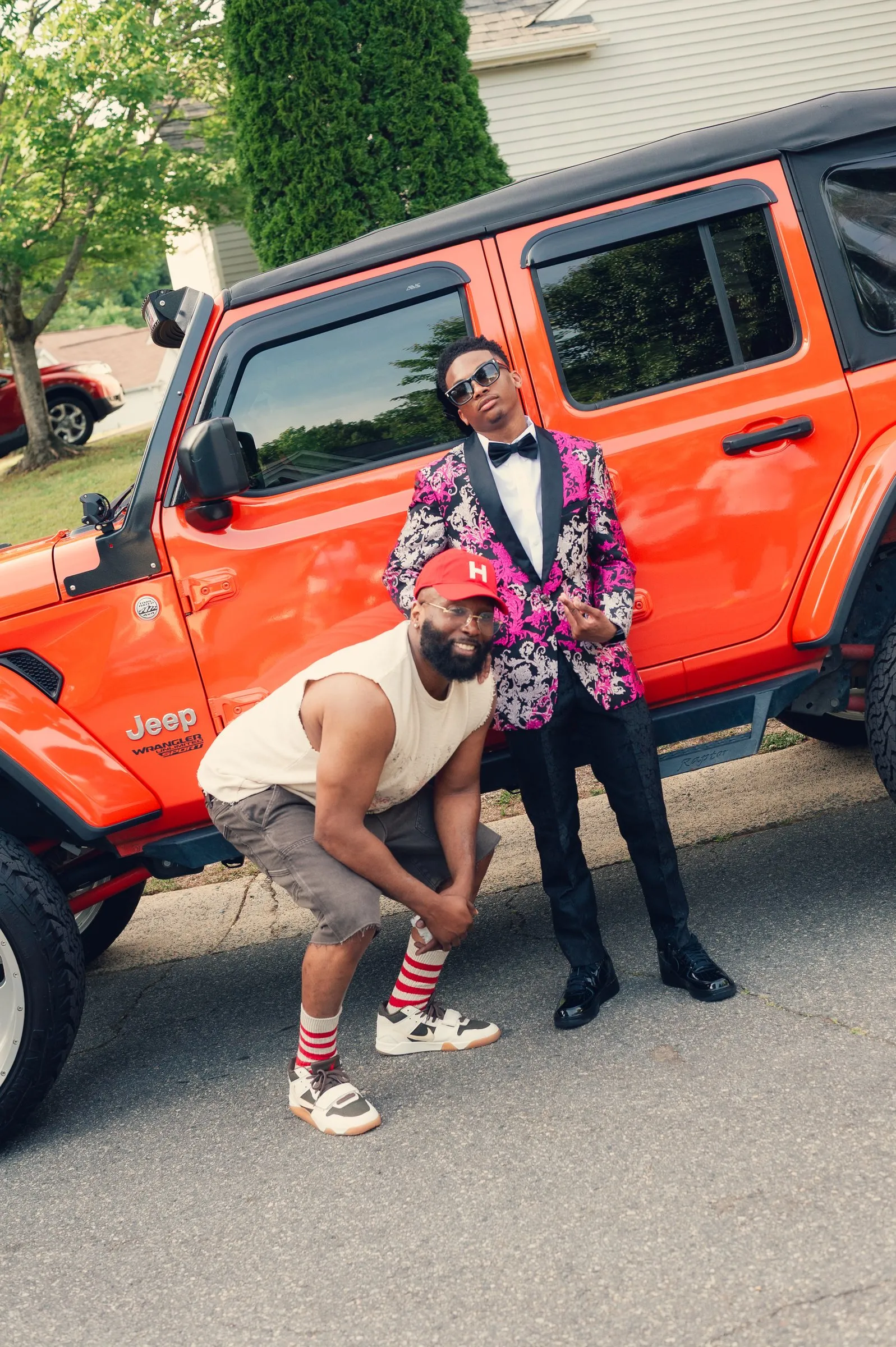 He poses in front of an orange Jeep Wrangler while a man in a red cap crouches beside him