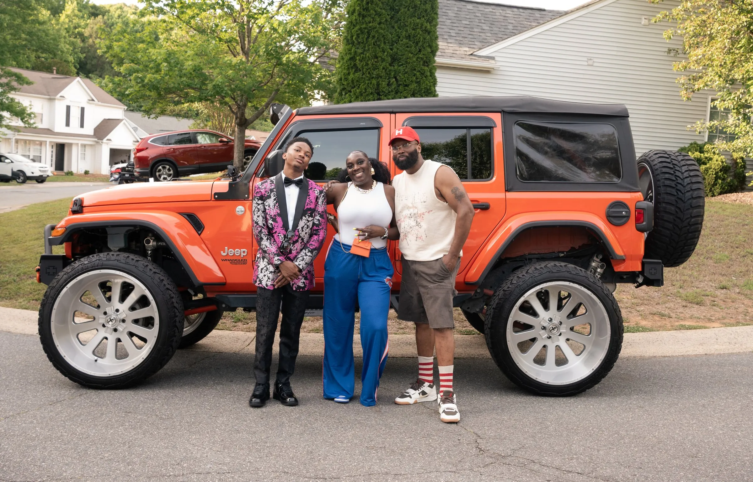 He stands with a woman in blue track pants and a man in a red cap beside the orange Jeep
