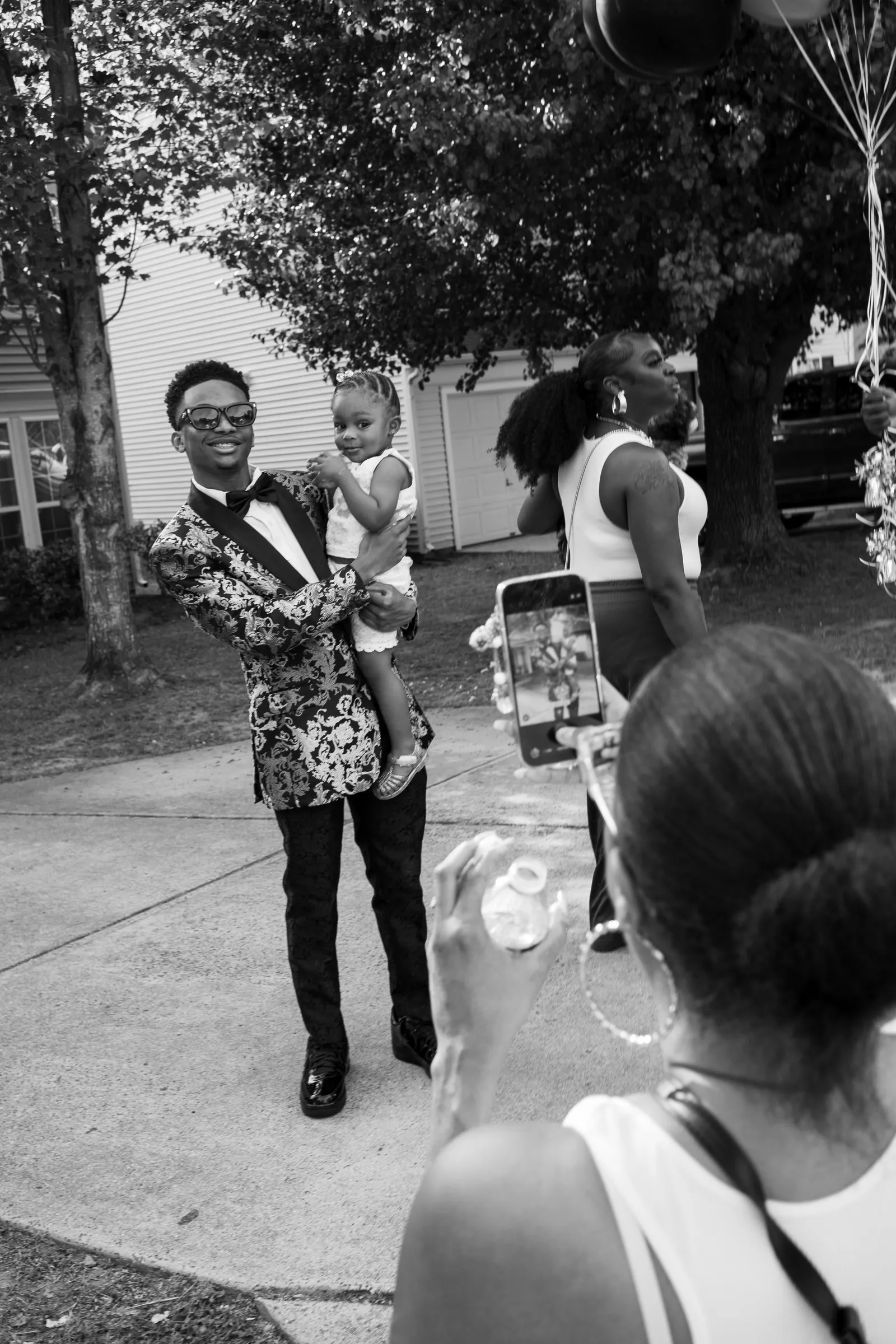 Black and white photograph of him carrying a toddler down the driveway past guests holding up phones