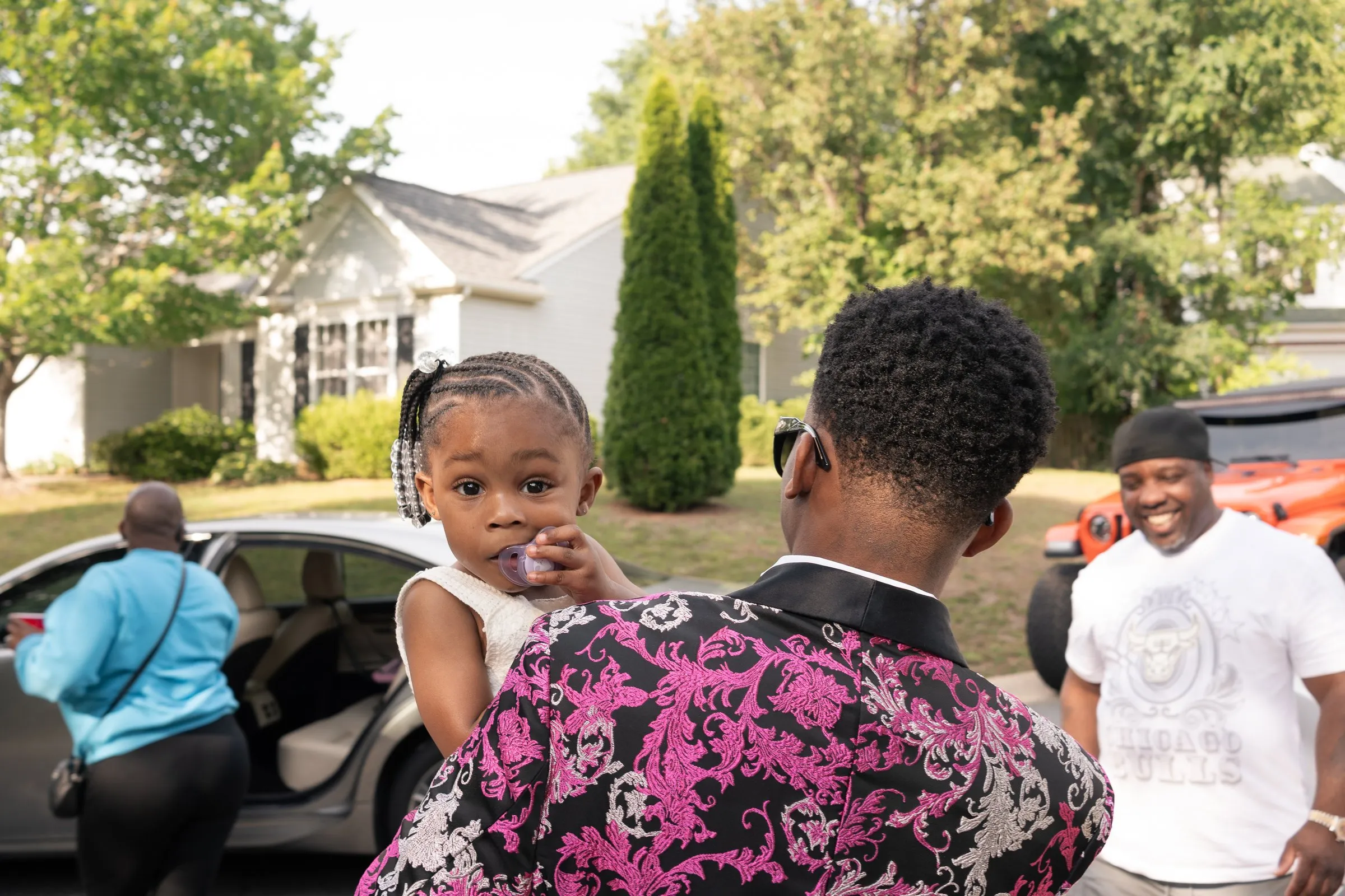 He holds a little girl on his shoulder as guests gather in the street
