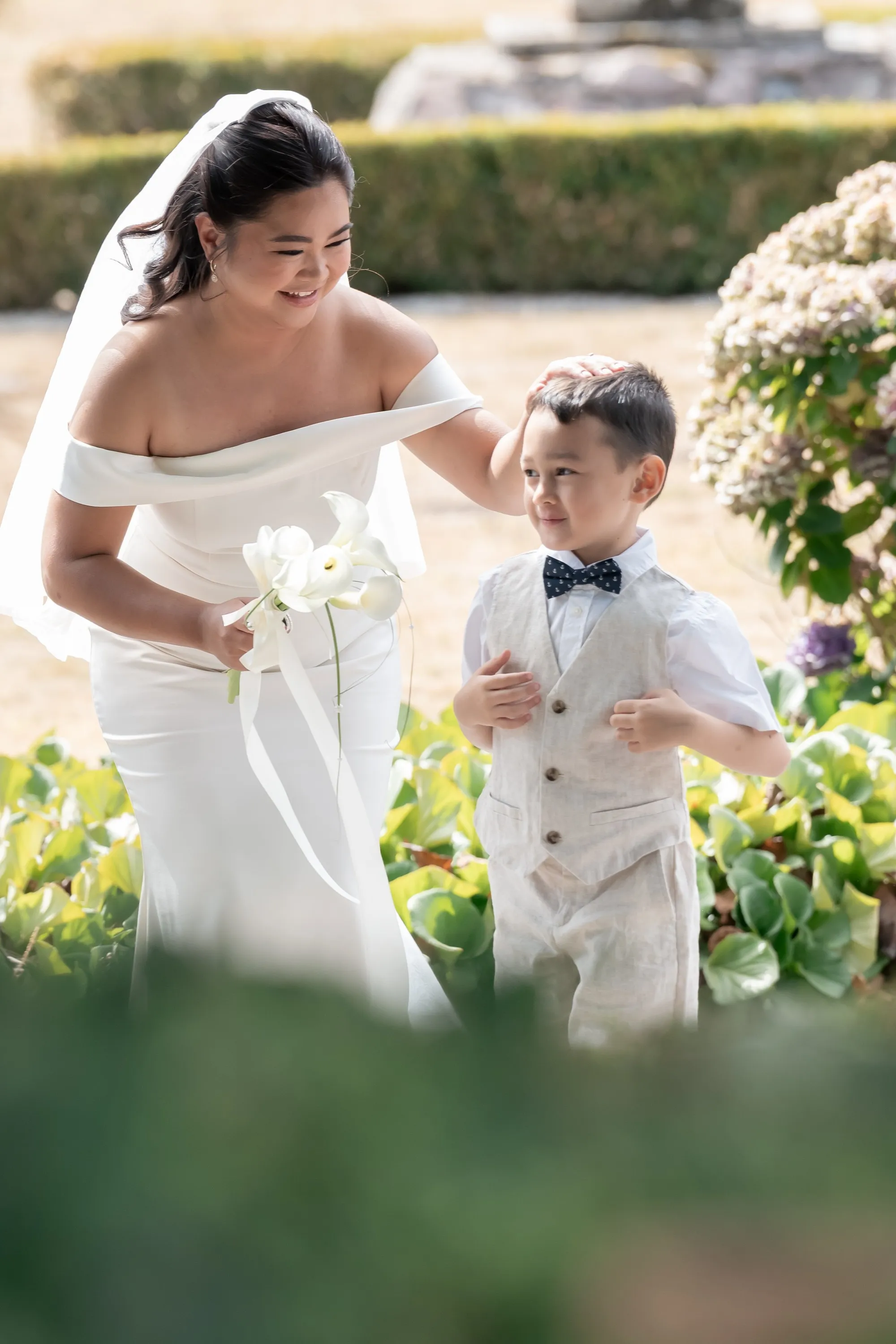 Bride in an off-the-shoulder gown walking through a garden with a young ring bearer in a linen vest and bow tie, Signature edit