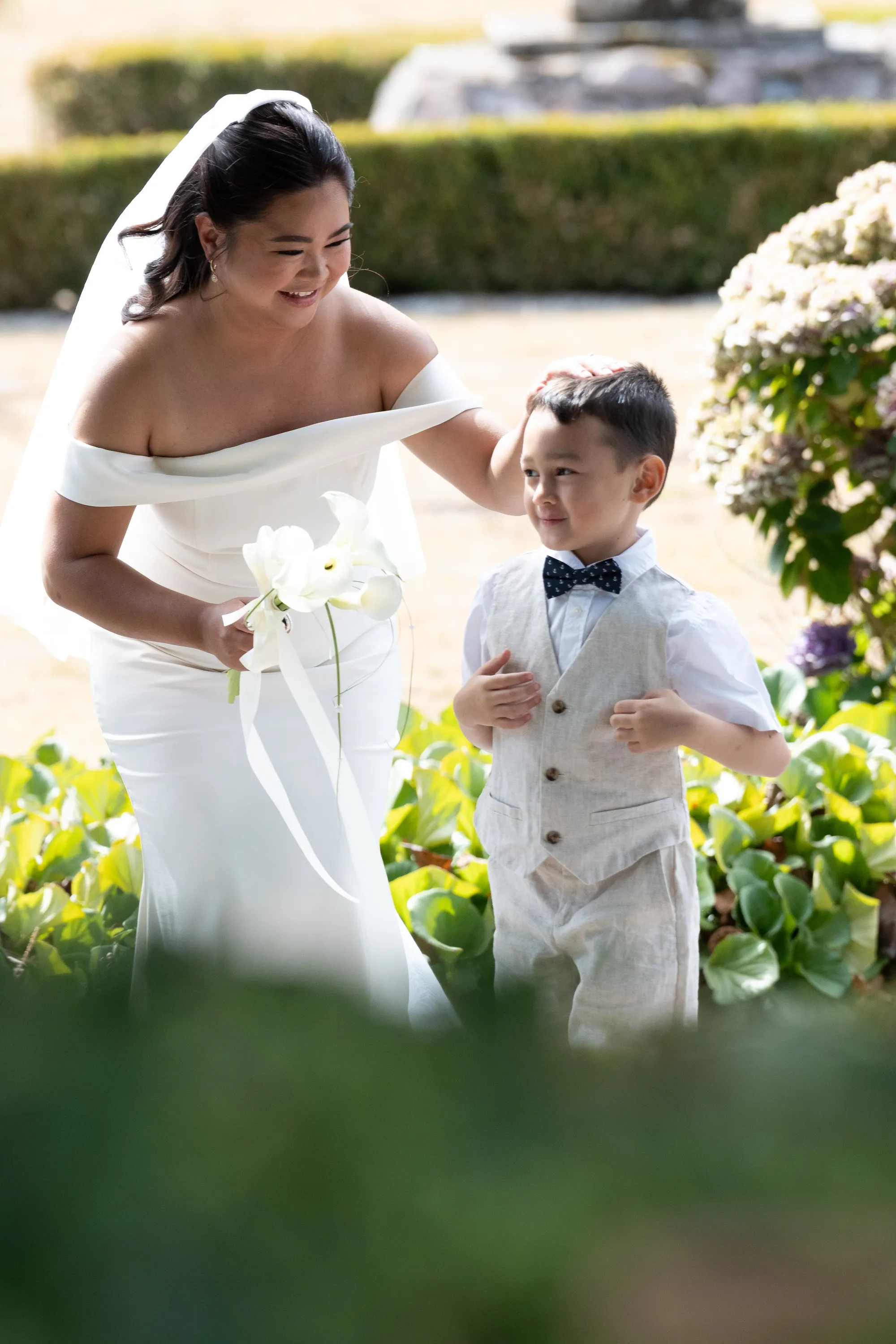 Bride in an off-the-shoulder gown walking through a garden with a young ring bearer in a linen vest and bow tie, Signature, straight out of the camera before editing