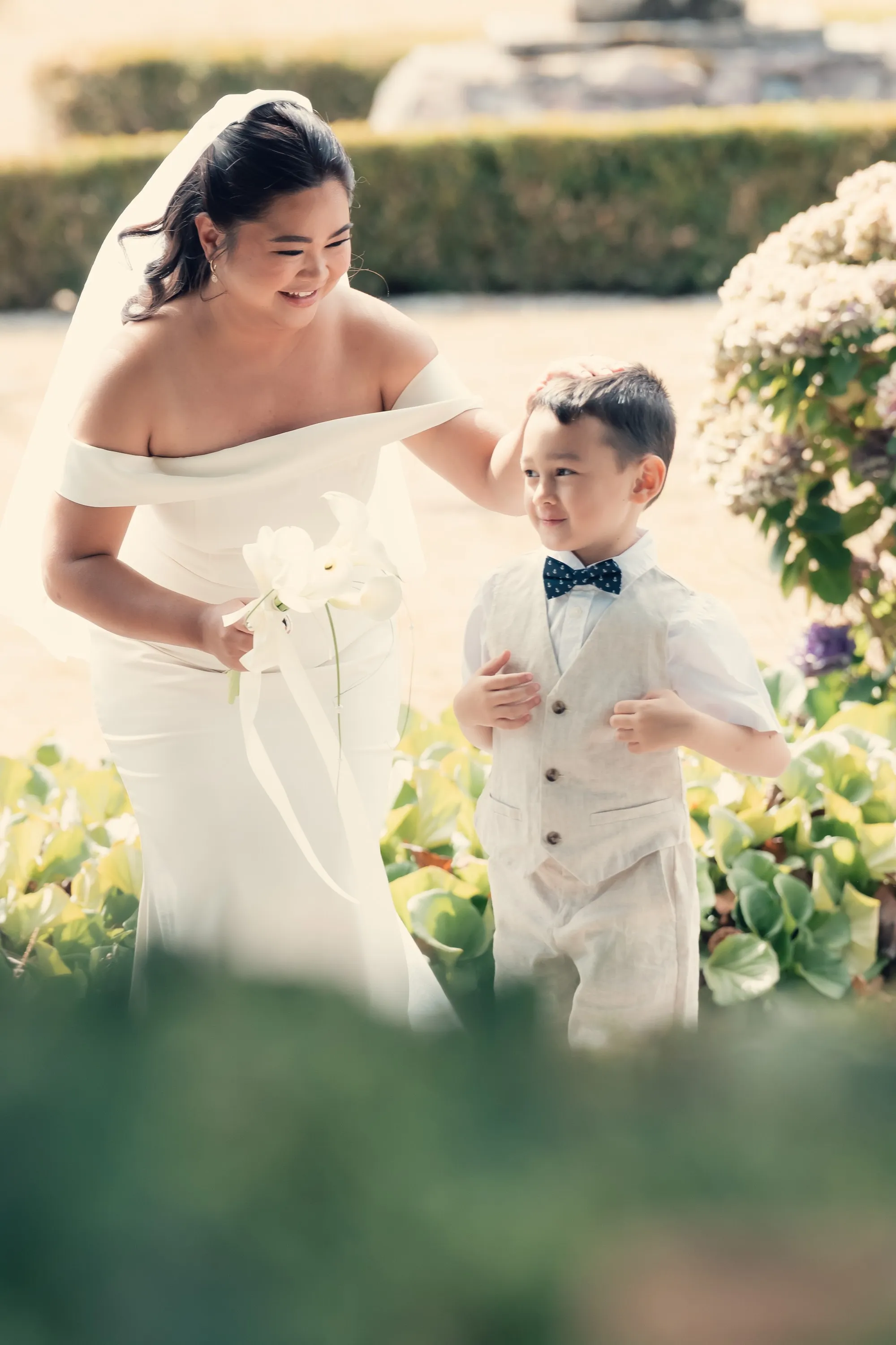 Bride in an off-the-shoulder gown walking through a garden with a young ring bearer in a linen vest and bow tie, Signature Film edit