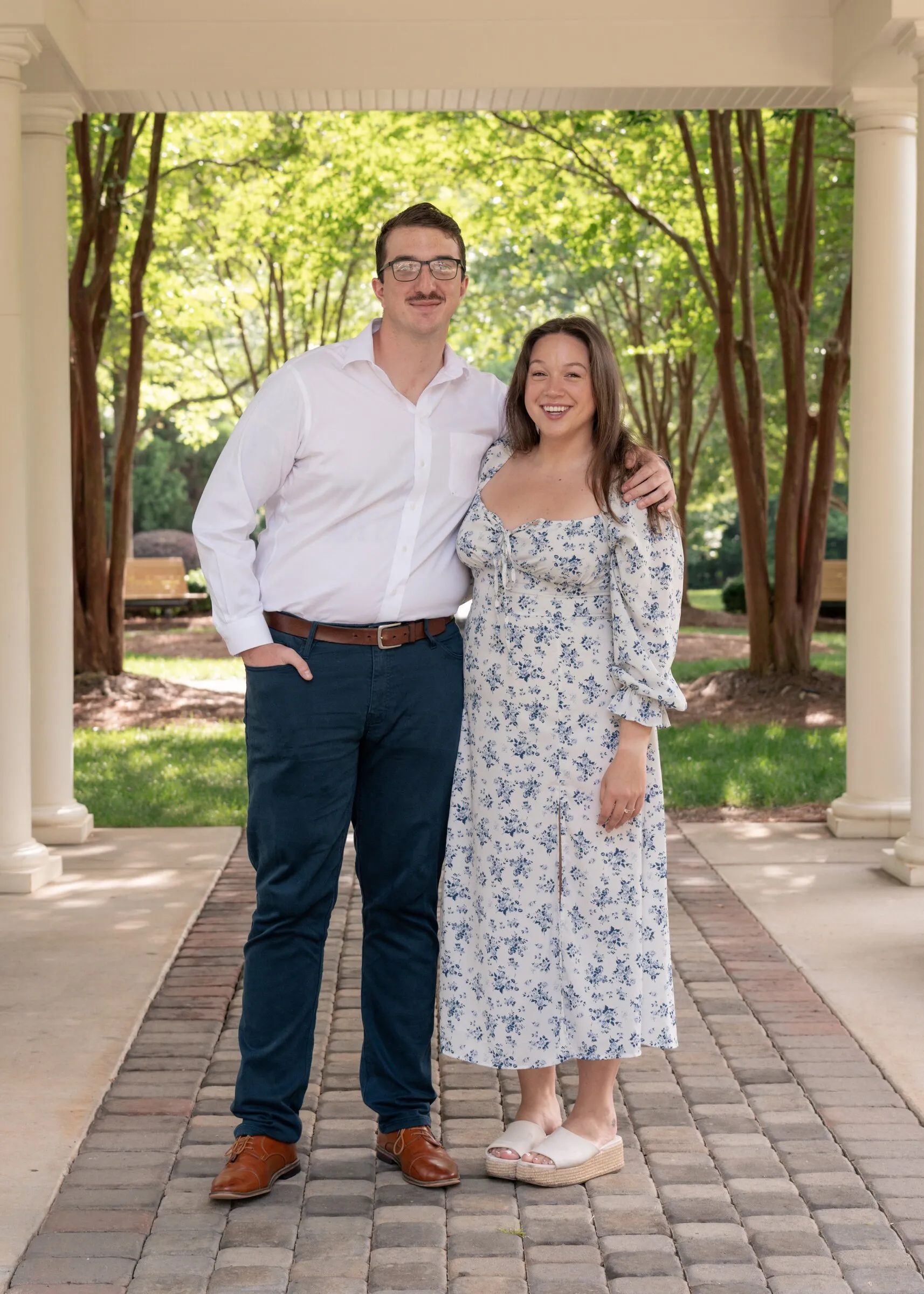 Spencer and Kayla standing together under a white columned pavilion at Millbridge, a brick path and crape myrtles behind them