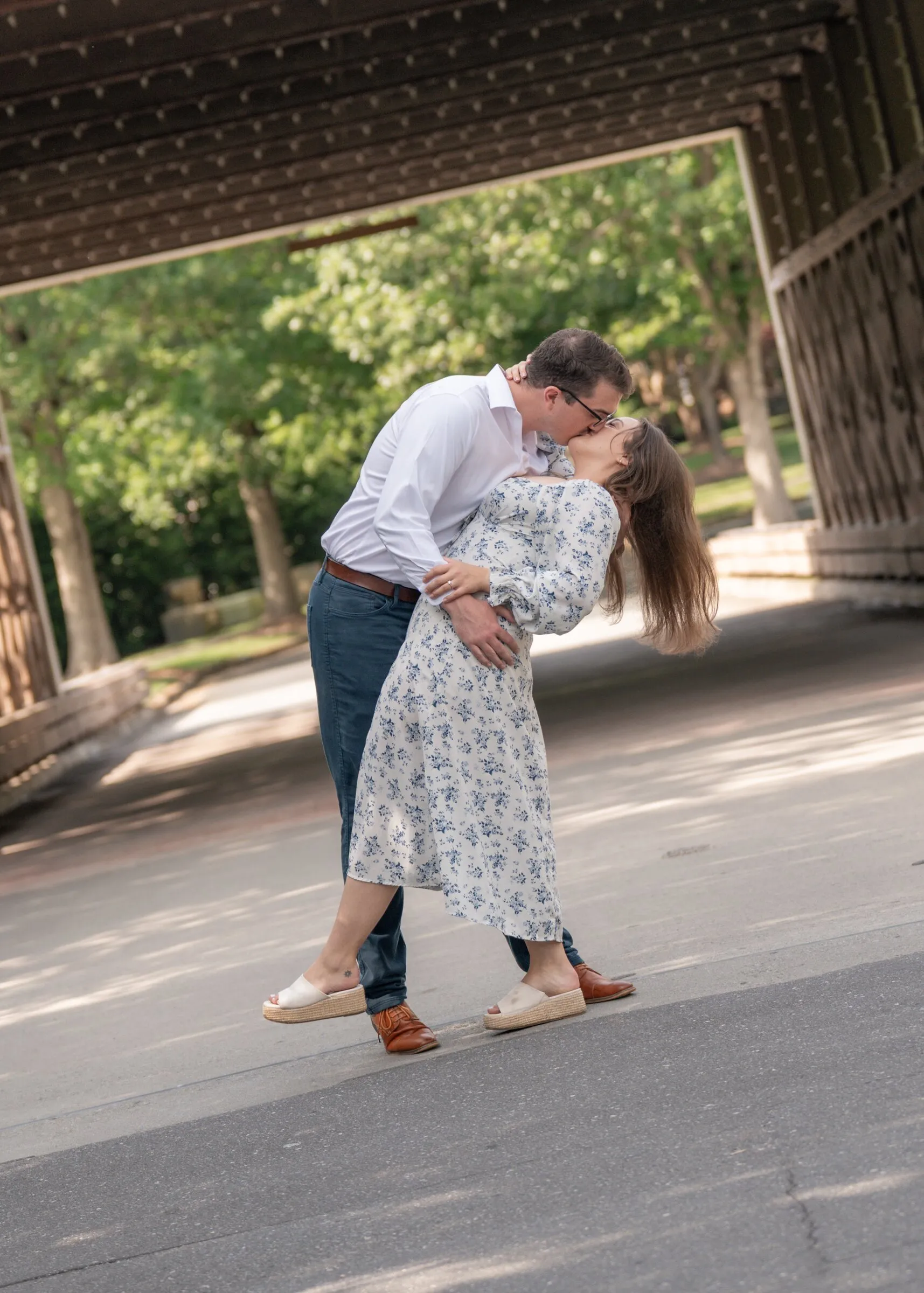 Spencer dipping Kayla for a kiss in the shade of the covered bridge, trees bright beyond the far end