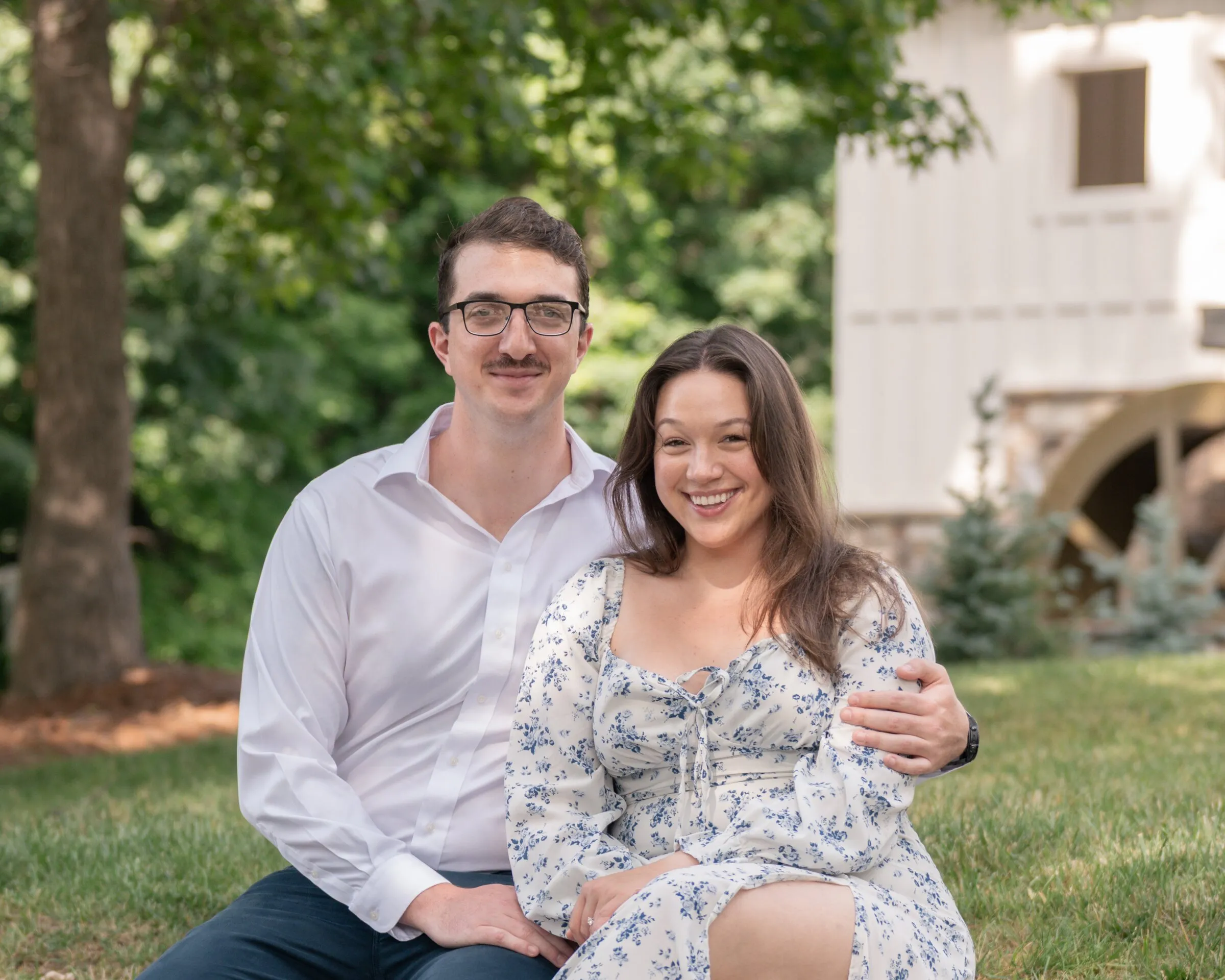 Spencer and Kayla seated on the lawn beside the white mill house, his arms around her