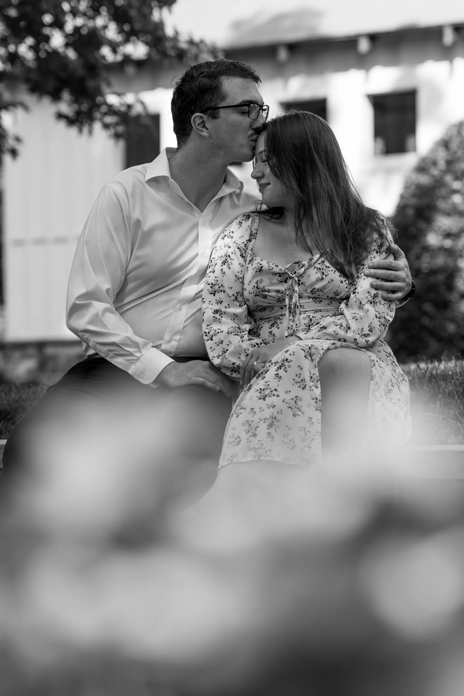 Black and white photograph of Spencer kissing Kayla's forehead as they sit beside the mill