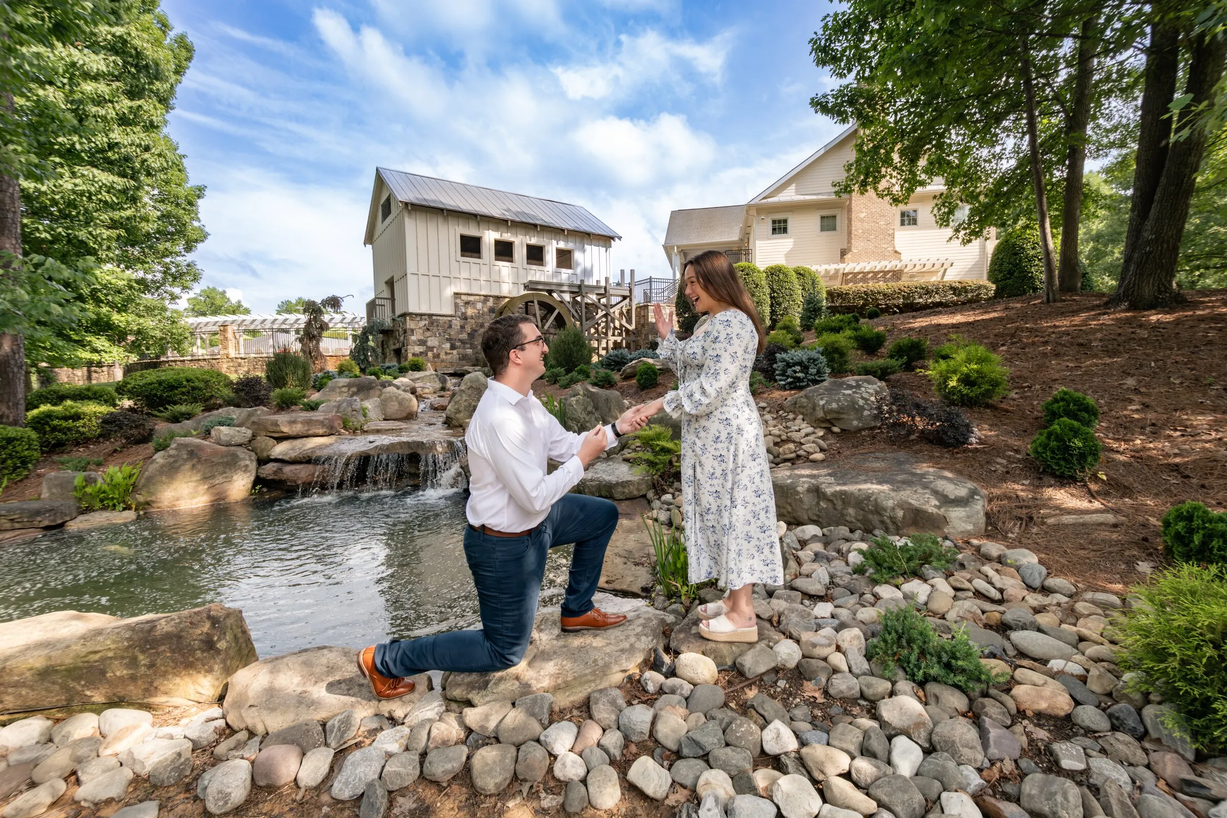 Spencer on one knee holding Kayla's hand on the rocks by the waterfall, the mill house and blue sky above