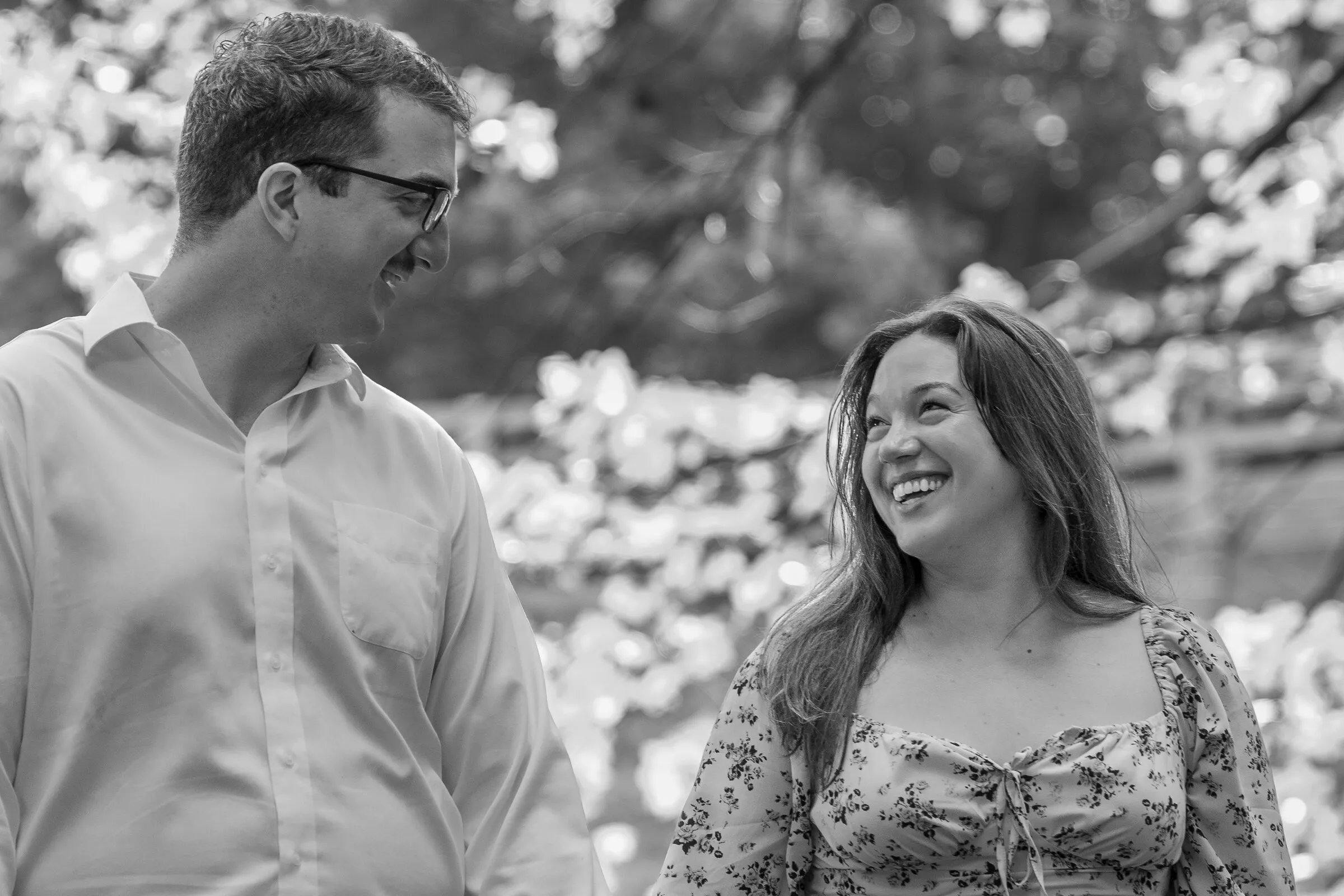 Black and white frame of Spencer and Kayla smiling at each other in front of sunlit leaves