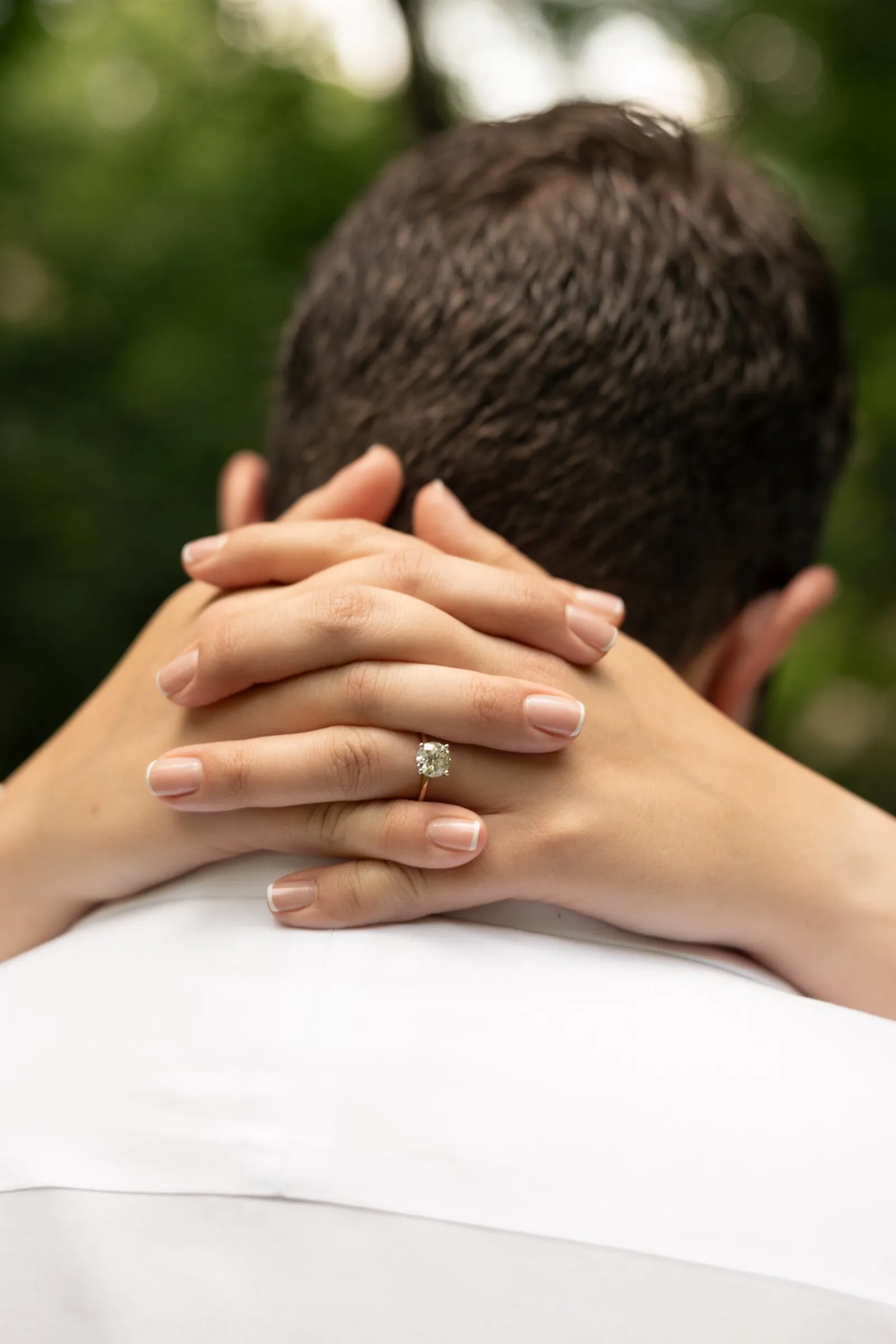 Kayla's hands clasped behind Spencer's neck, her engagement ring in view against dark green trees