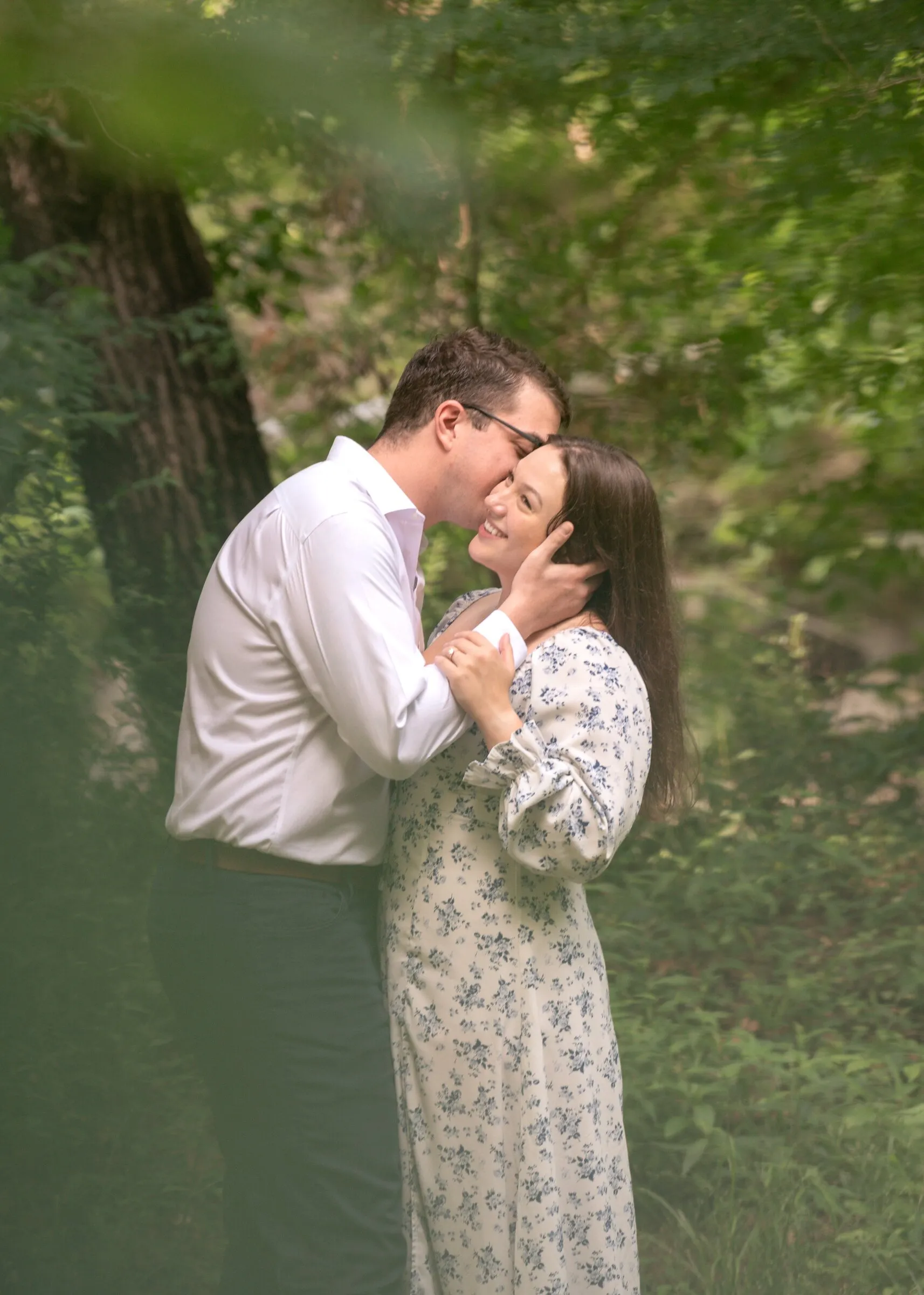 Spencer kissing Kayla's cheek among the trees, leaves soft in the foreground