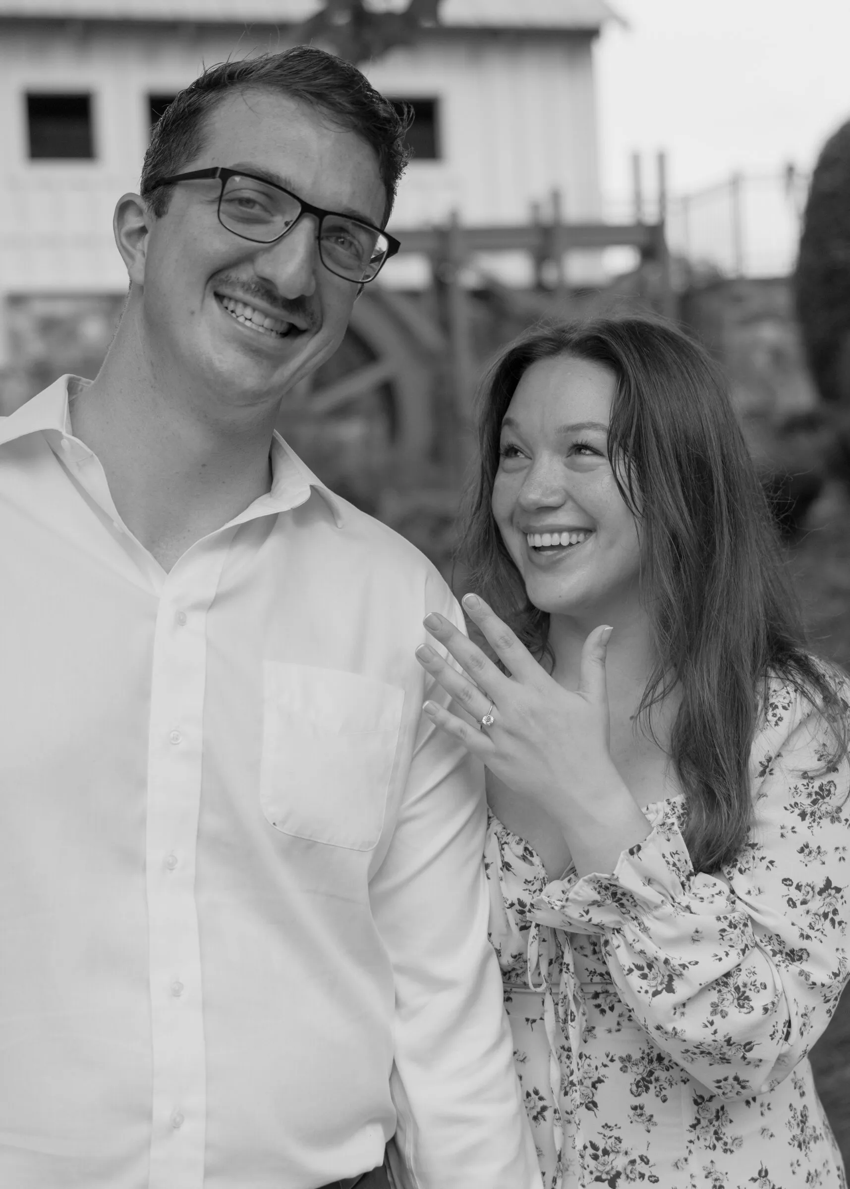 Black and white photograph of Spencer grinning at the camera as Kayla admires her ring
