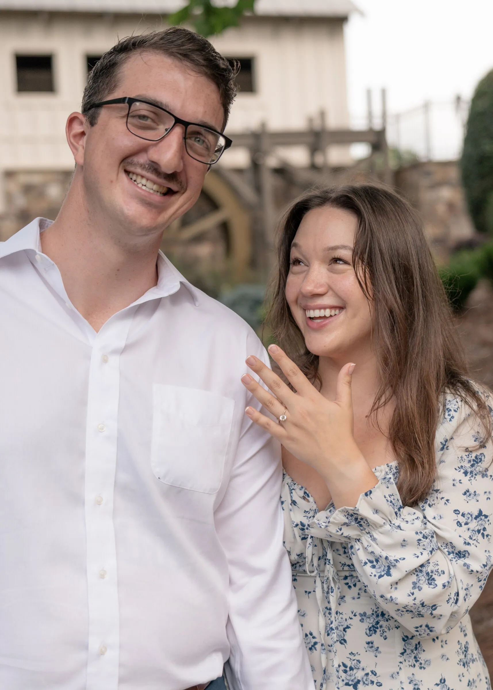 Spencer grinning at the camera while Kayla smiles at her engagement ring in front of the mill