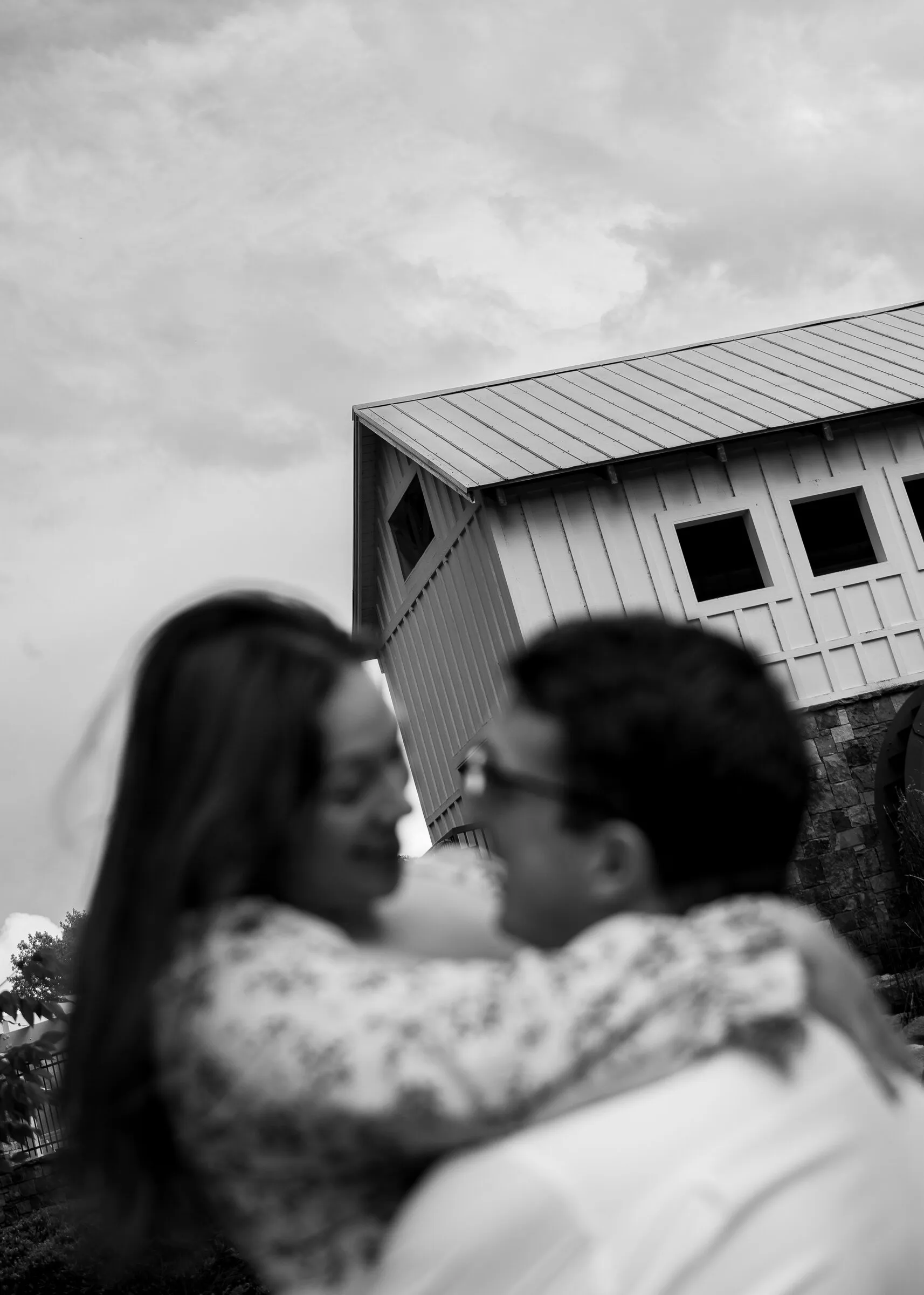Black and white frame of Spencer and Kayla smiling face to face beneath the mill's roofline