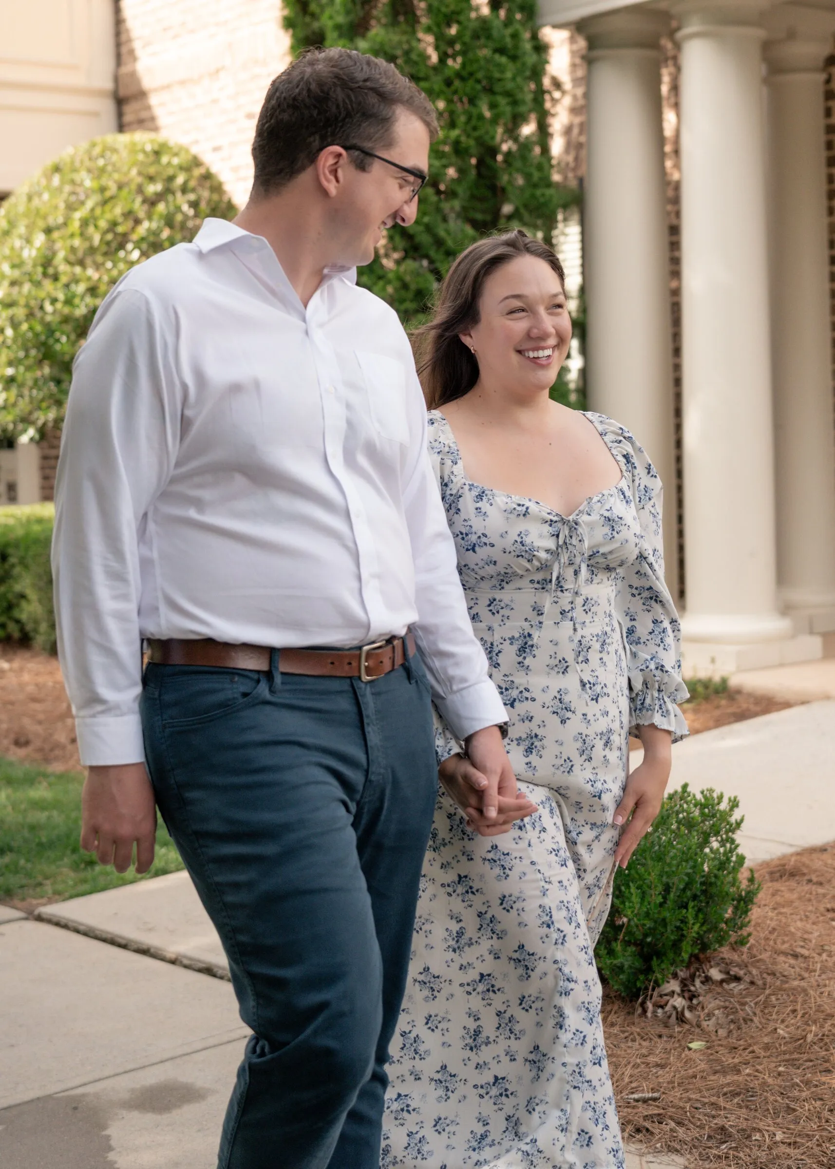 Spencer and Kayla walking hand in hand past a columned porch, both laughing