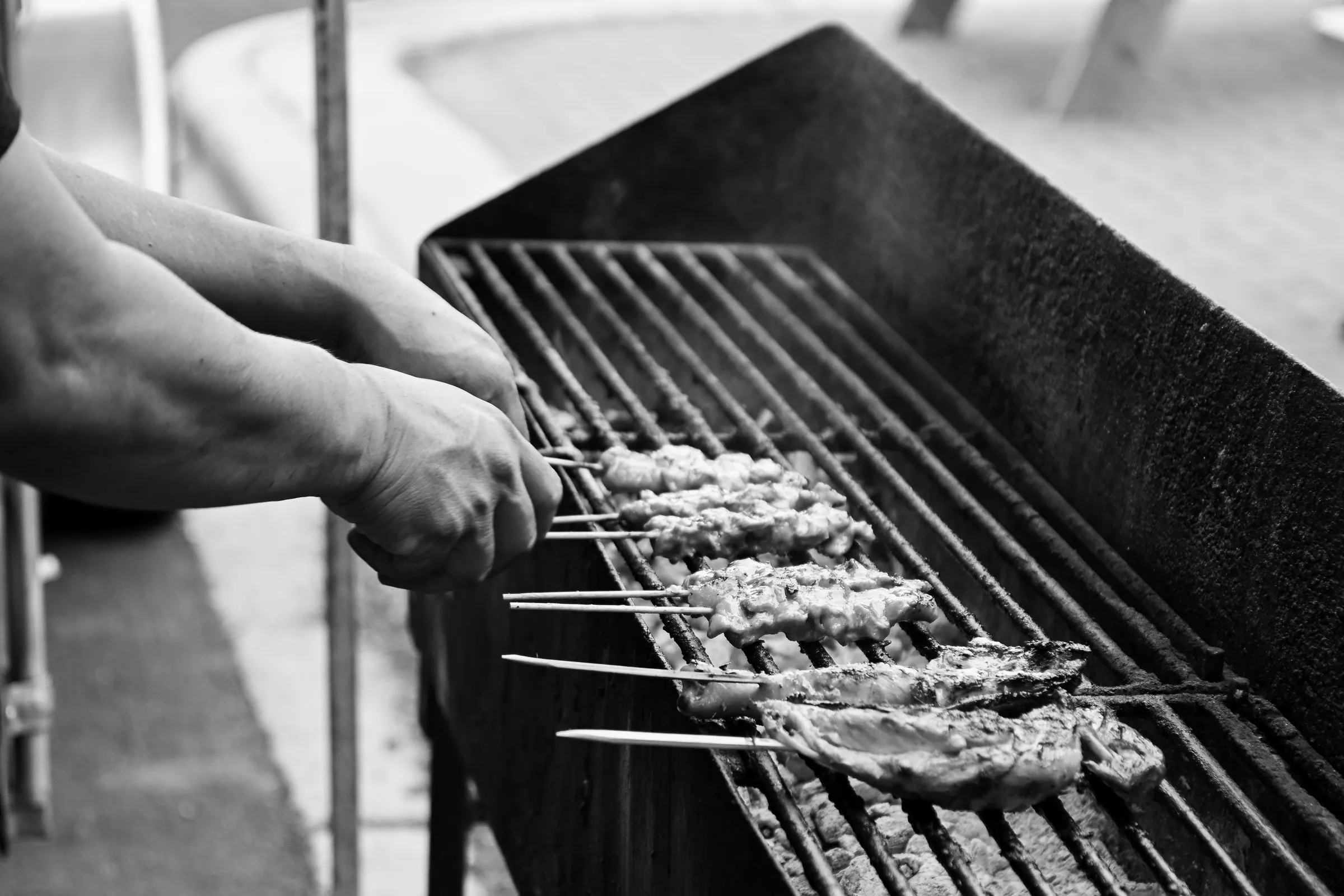 Black and white photograph of hands turning a row of chicken skewers over the coals