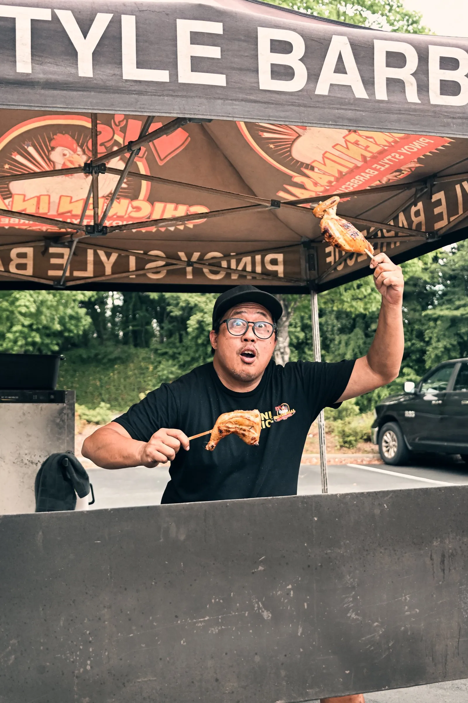 The cook holds a chicken inasal skewer high under the Pinoy Style Barbecue tent