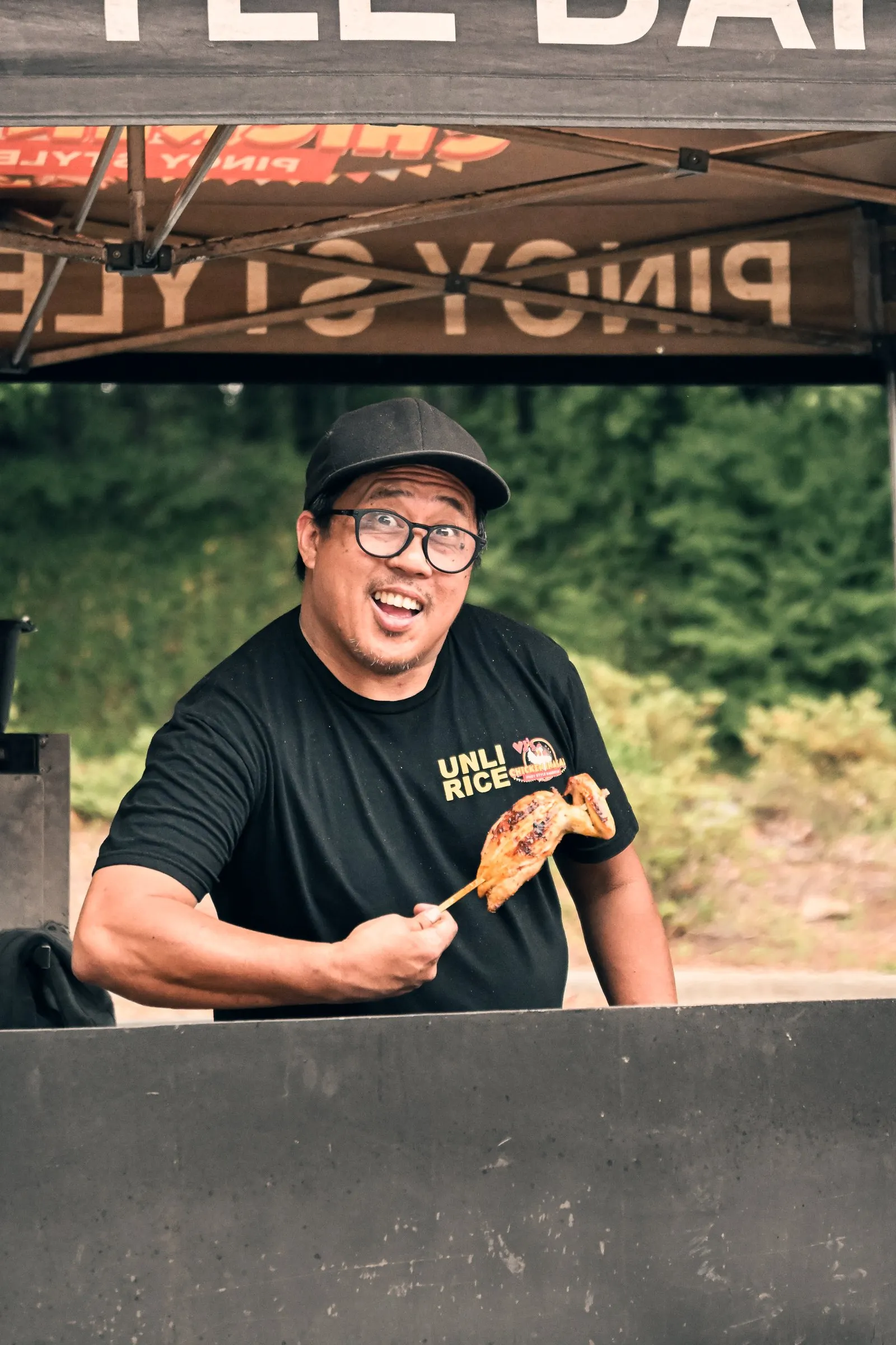 The cook laughs while holding a fresh skewer of chicken inasal behind the grill