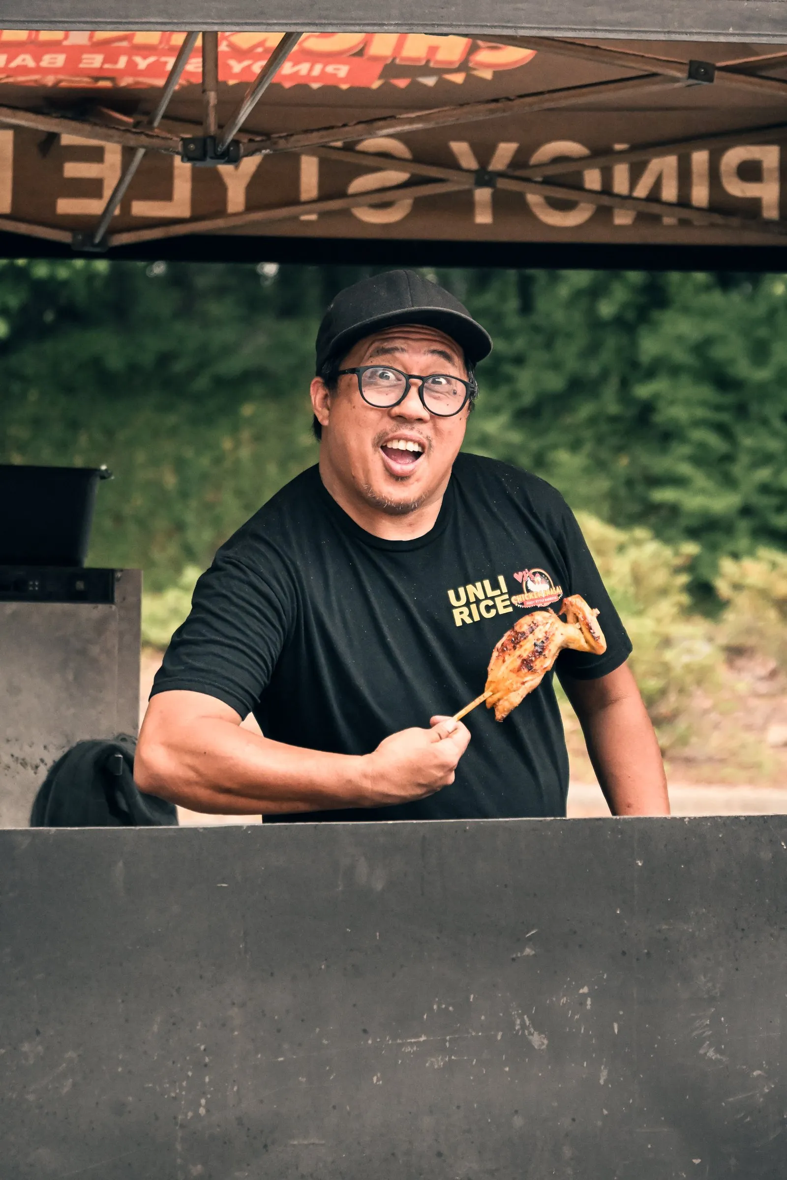 The cook raises a grilled chicken skewer under the VJ's Chicken Inasal tent