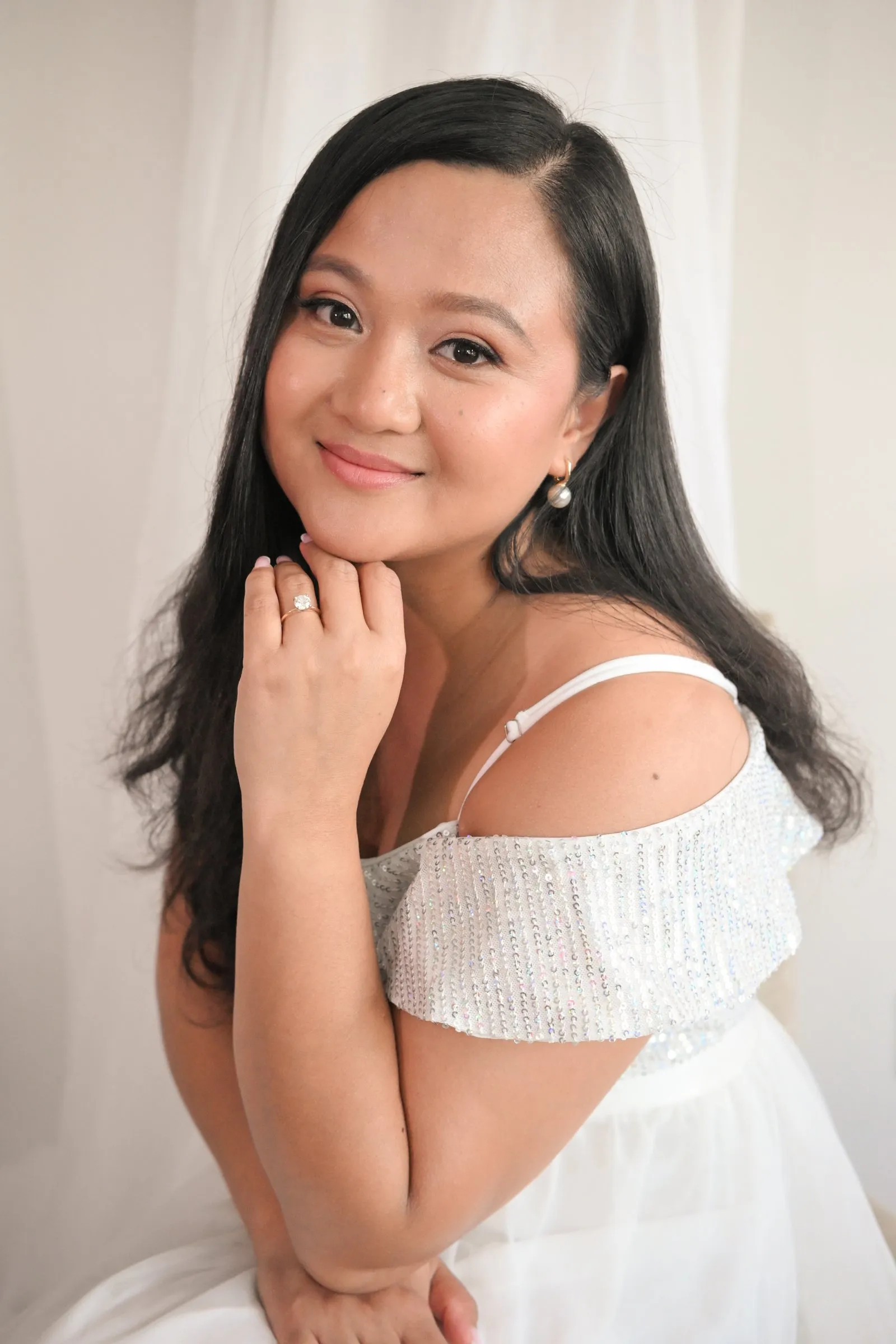 Bridal portrait of Winnie in an off the shoulder white sequin dress and pearl earrings, her chin resting on her hand and her engagement ring showing