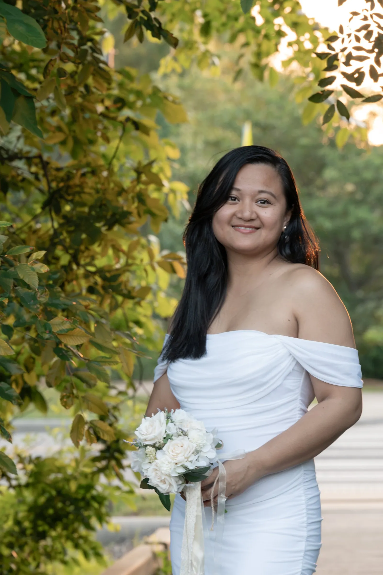 Winnie smiling in an off the shoulder white gown, holding a bouquet of cream roses under a canopy of green leaves