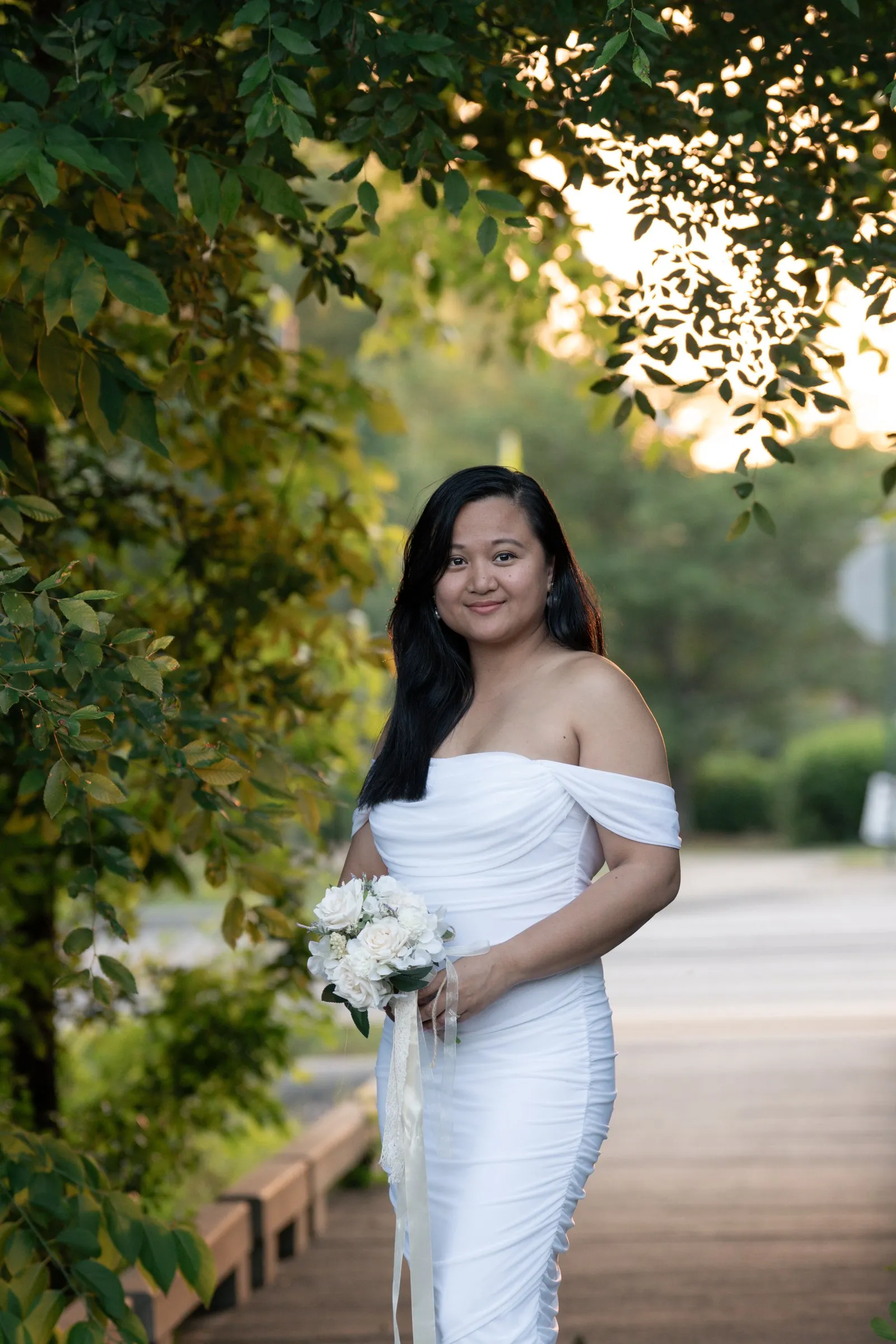 Winnie on a wooden boardwalk path in her white gown, bouquet ribbons trailing, with the sun low through the trees
