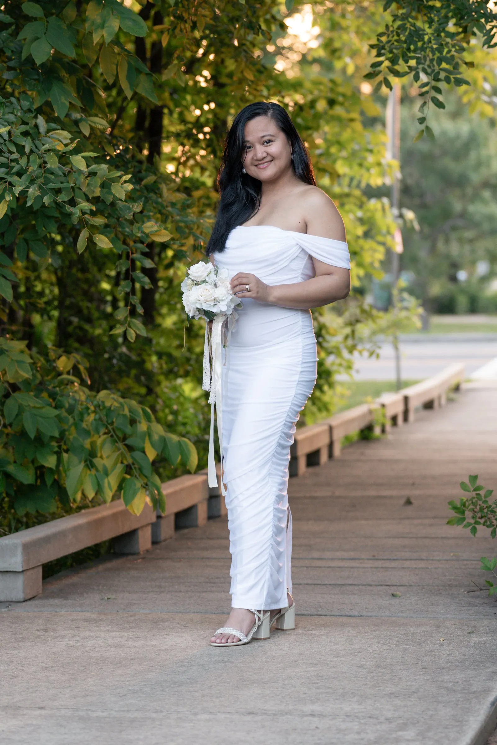 Full length portrait of Winnie on a boardwalk lined with greenery, her fitted ruched white gown reaching her sandals