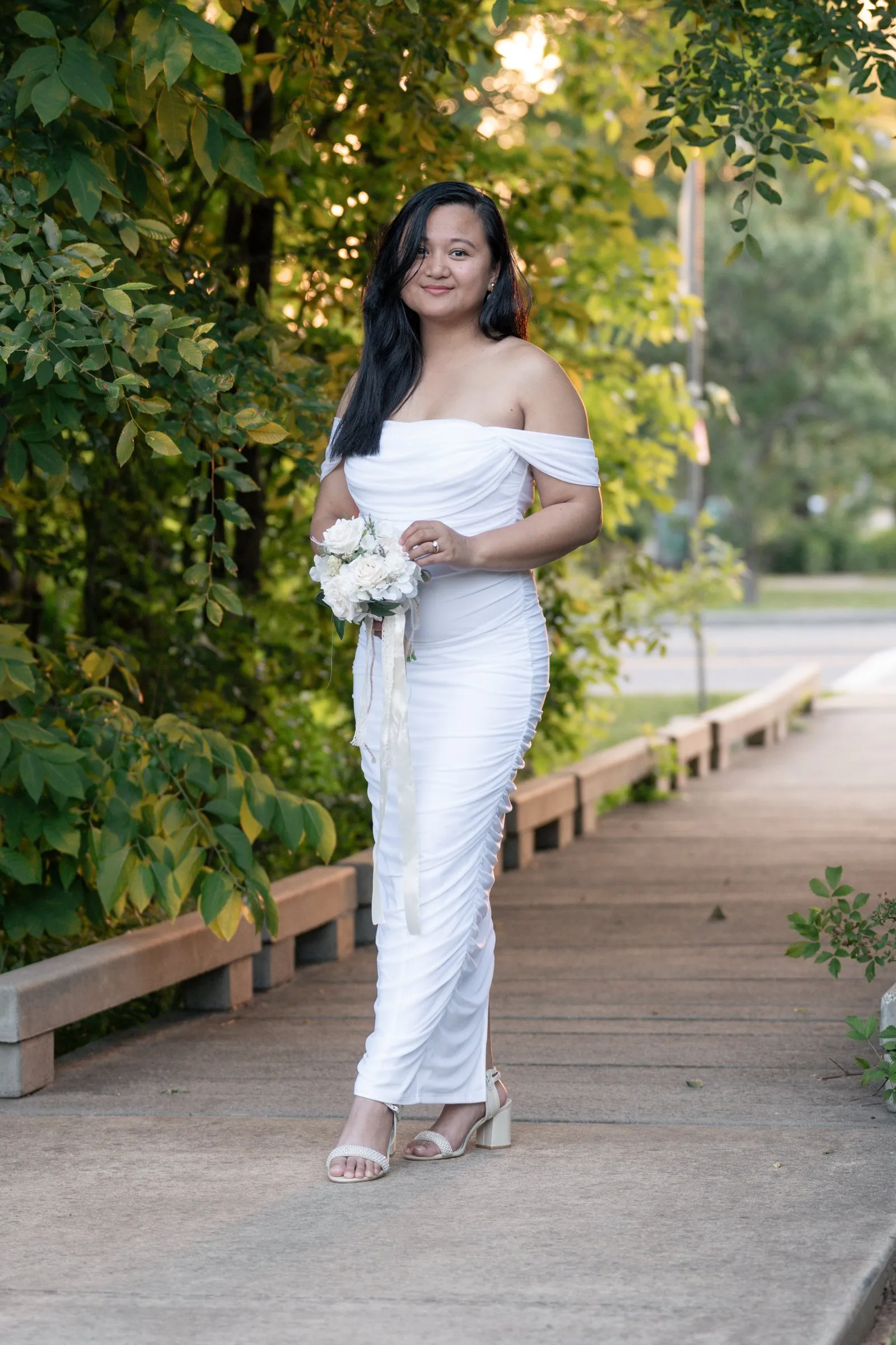 Winnie tilting her head and smiling on the boardwalk path, long ribbons falling from her bouquet