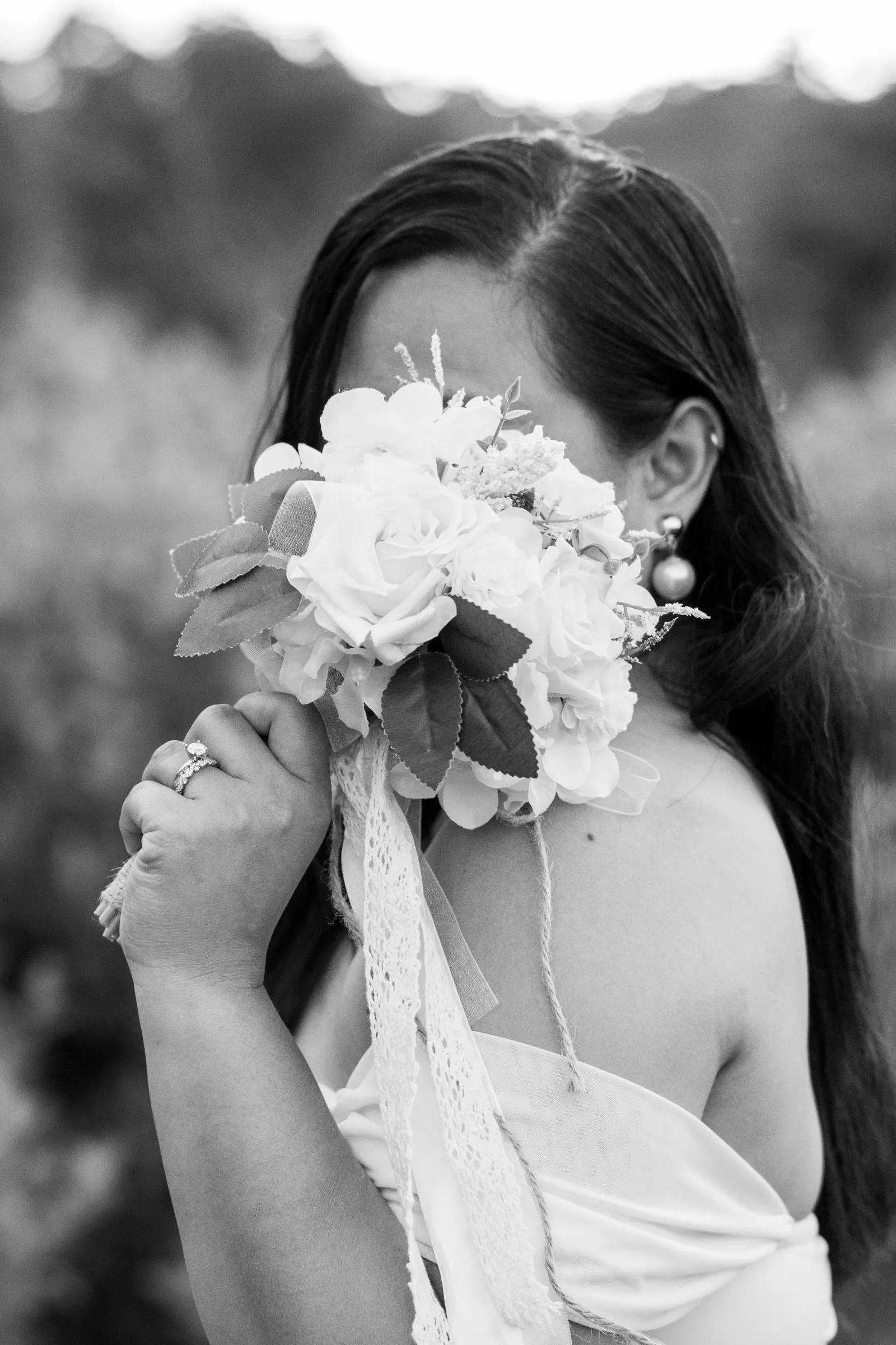 Black and white portrait of Winnie holding her bouquet up to cover her face, lace ribbons hanging from the stems