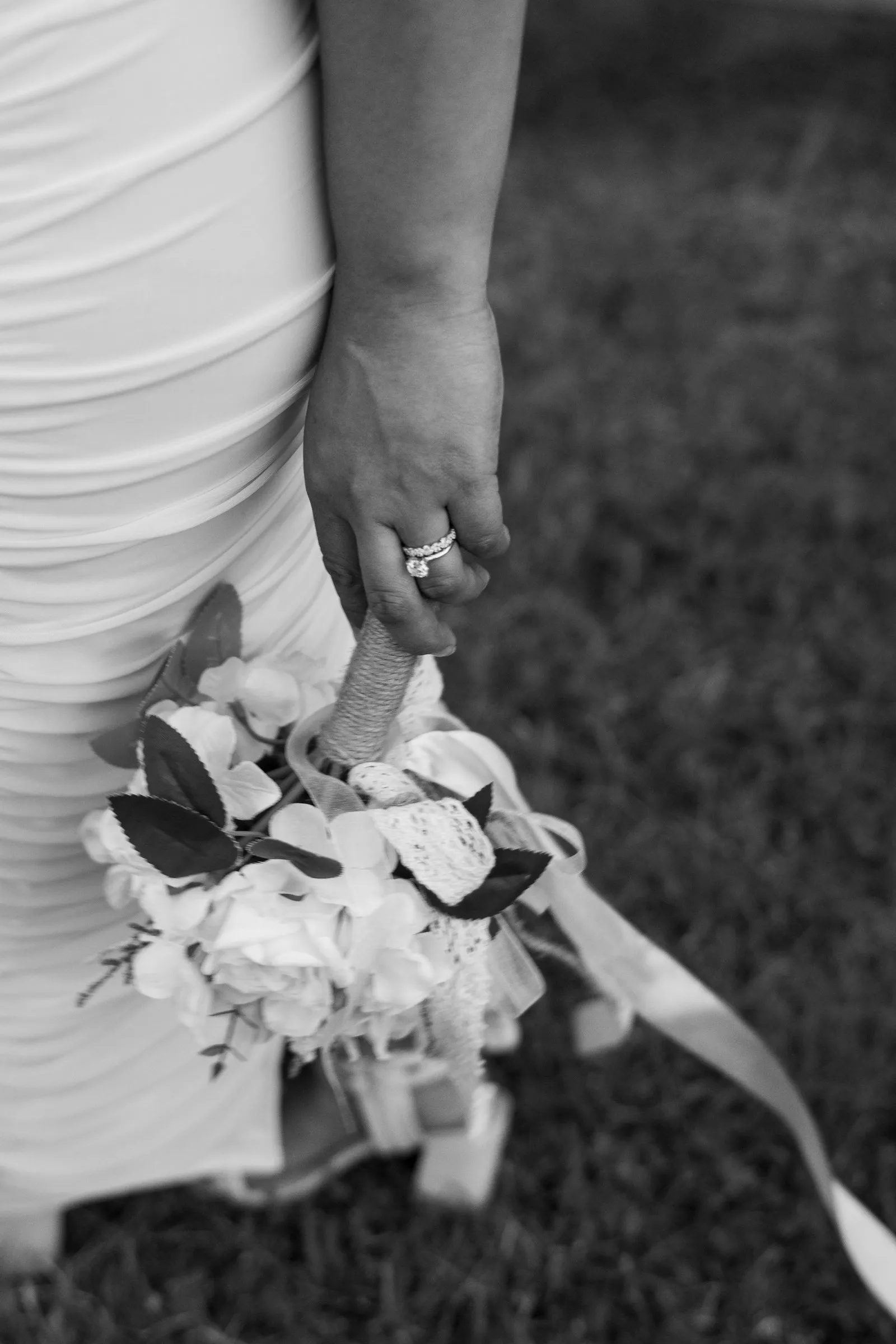 Black and white version of the same frame: Winnie's bouquet hanging at her side with ribbons drifting past her sandals