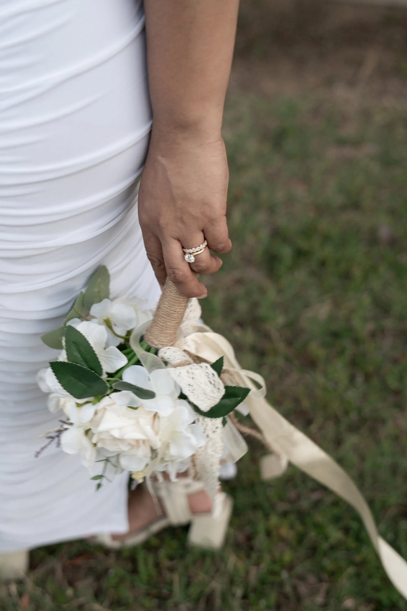 Winnie's hand at her side holding the bouquet by its twine wrapped stems, ivory ribbons and lace trailing into the grass