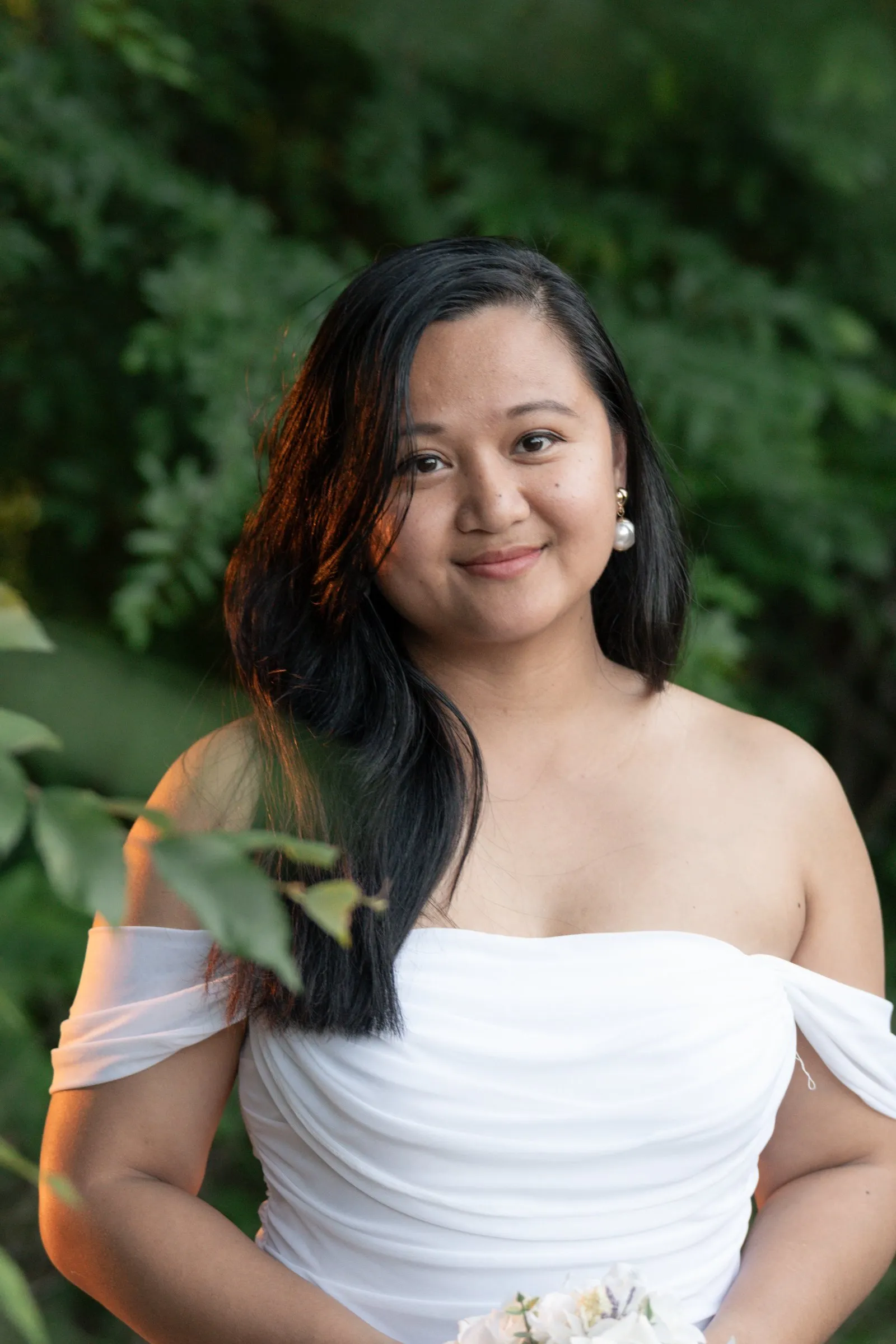 Close portrait of Winnie smiling softly in pearl drop earrings, warm light in her dark hair against deep green leaves